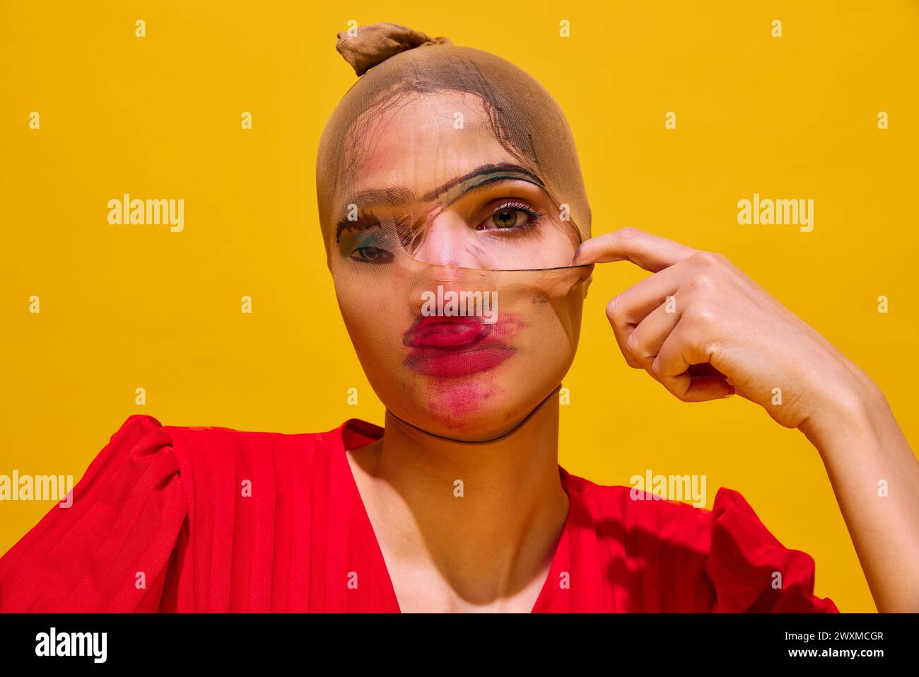 Young woman with stocking over head with smudged lipstick makeup against yellow background. Close-up. Secrets Stock Photo