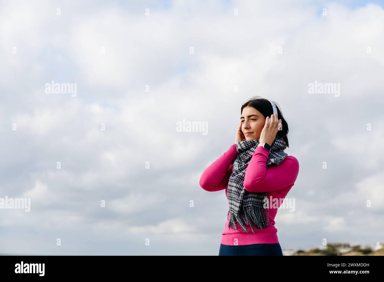 Woman in pink pullover and grey scarf listening music against sky Stock Photo