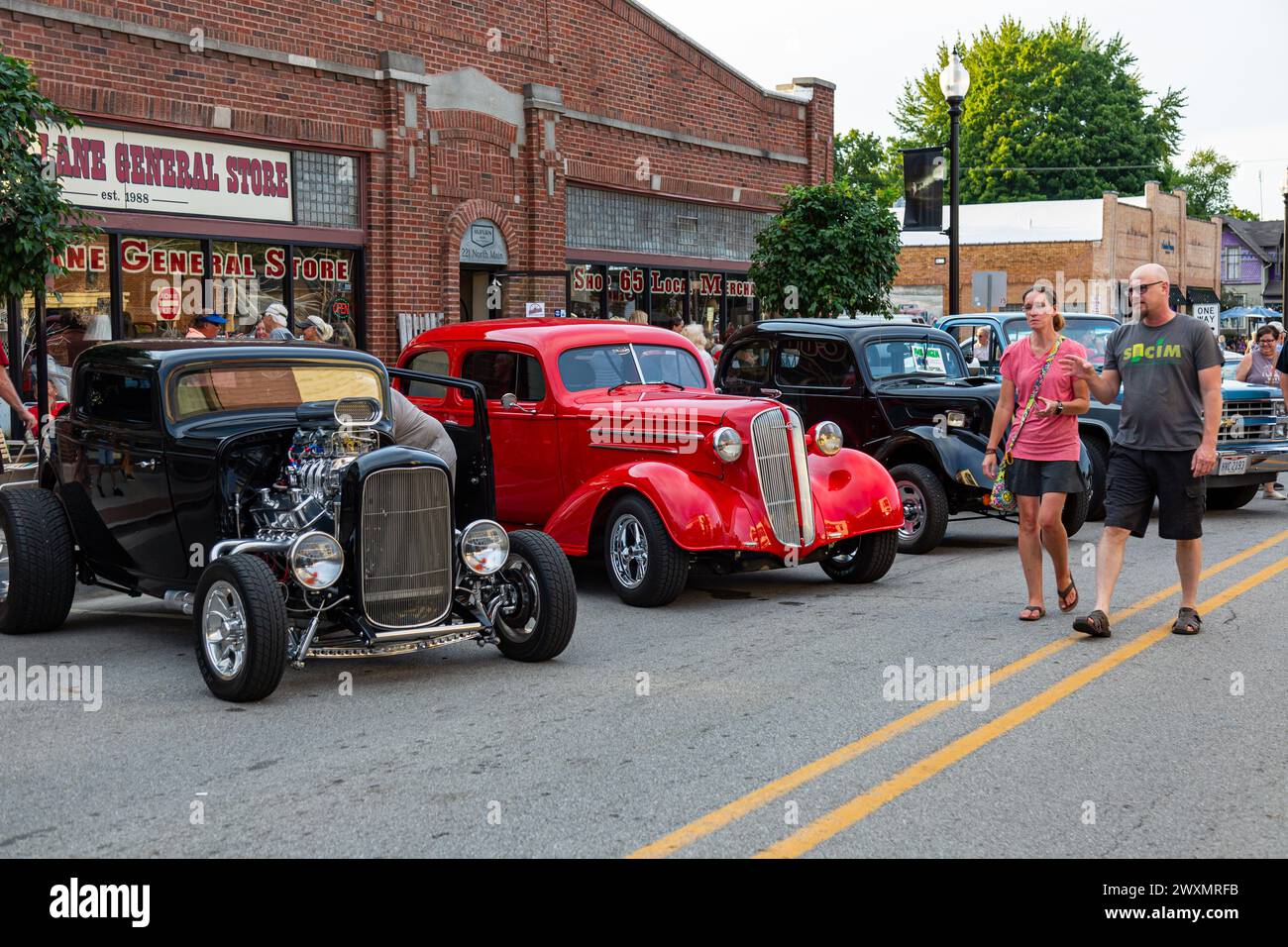 A couple walks past a red 1936 Chevrolet & a black 1932 Ford hotrod on display in front of the Country Lane General Store at a car show in Auburn, Ind. Stock Photo