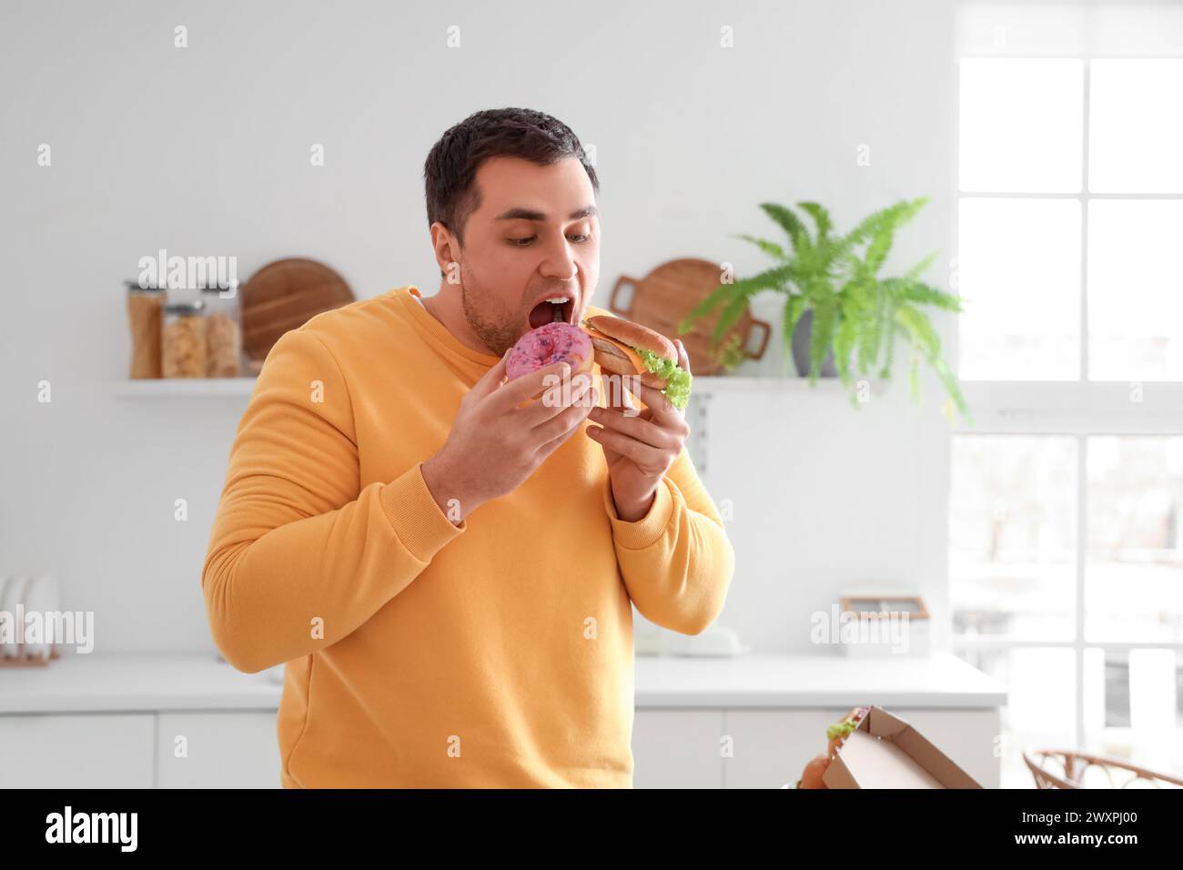 Young man eating burger and doughnut in kitchen. Overeating concept Stock Photo