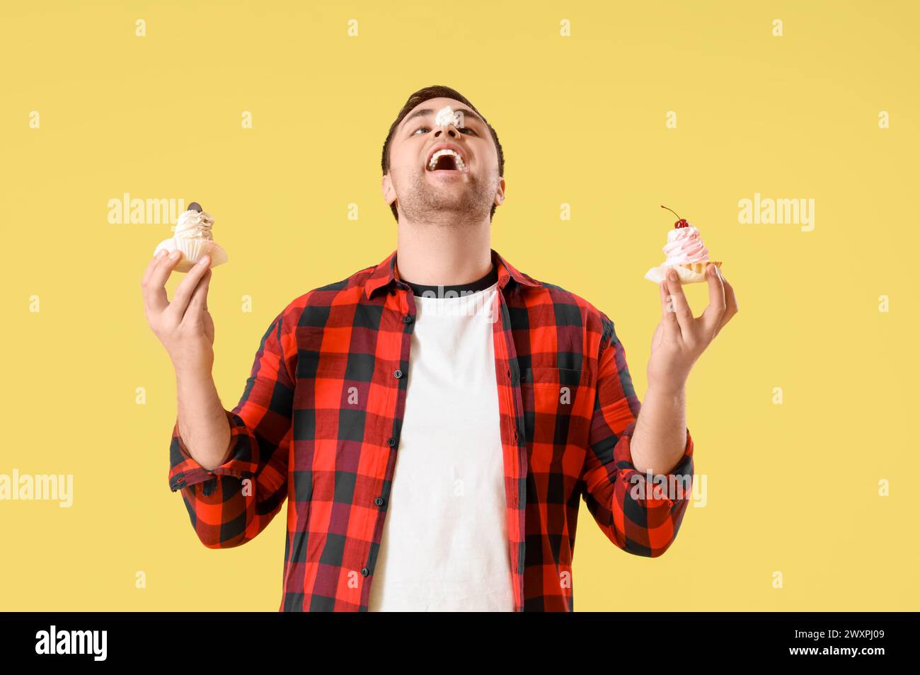 Young man with cream on his nose and cakes against yellow background. Overeating concept Stock Photo