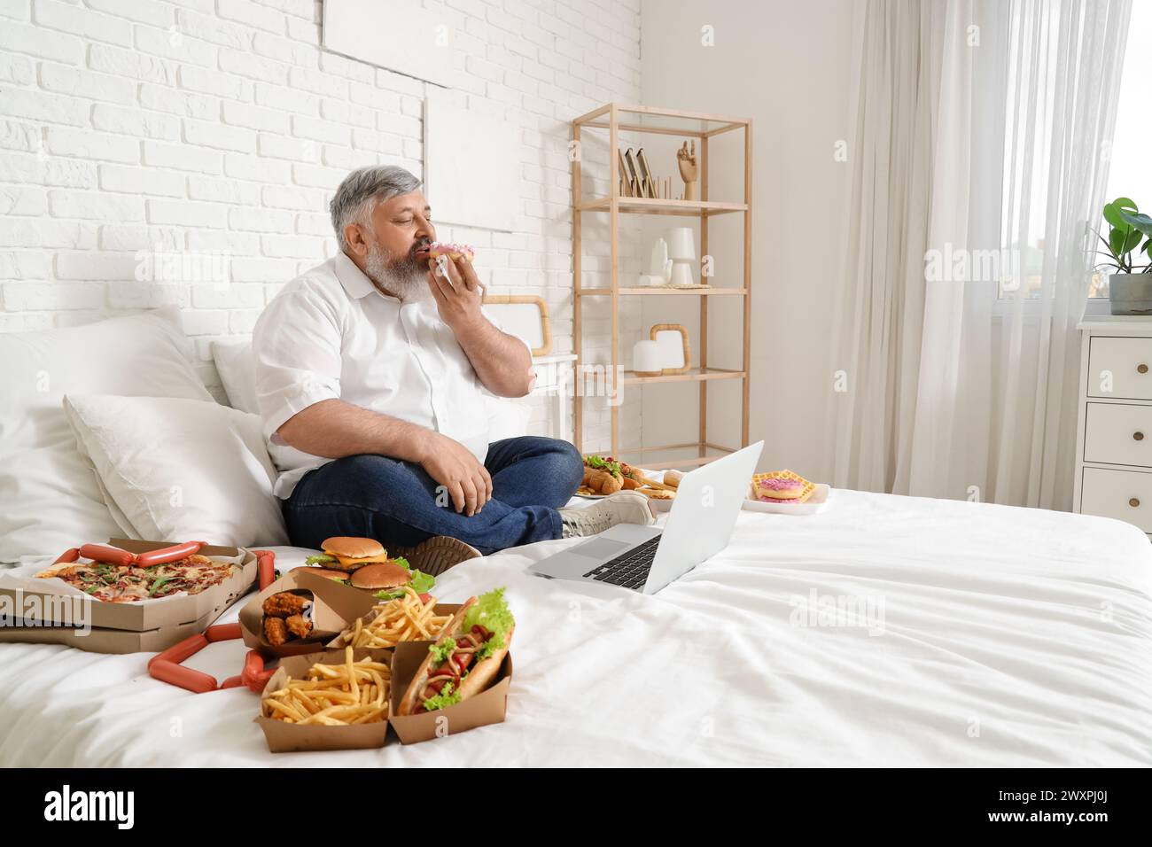 Overweight mature man with unhealthy food and laptop sitting in bedroom. Overeating concept Stock Photo