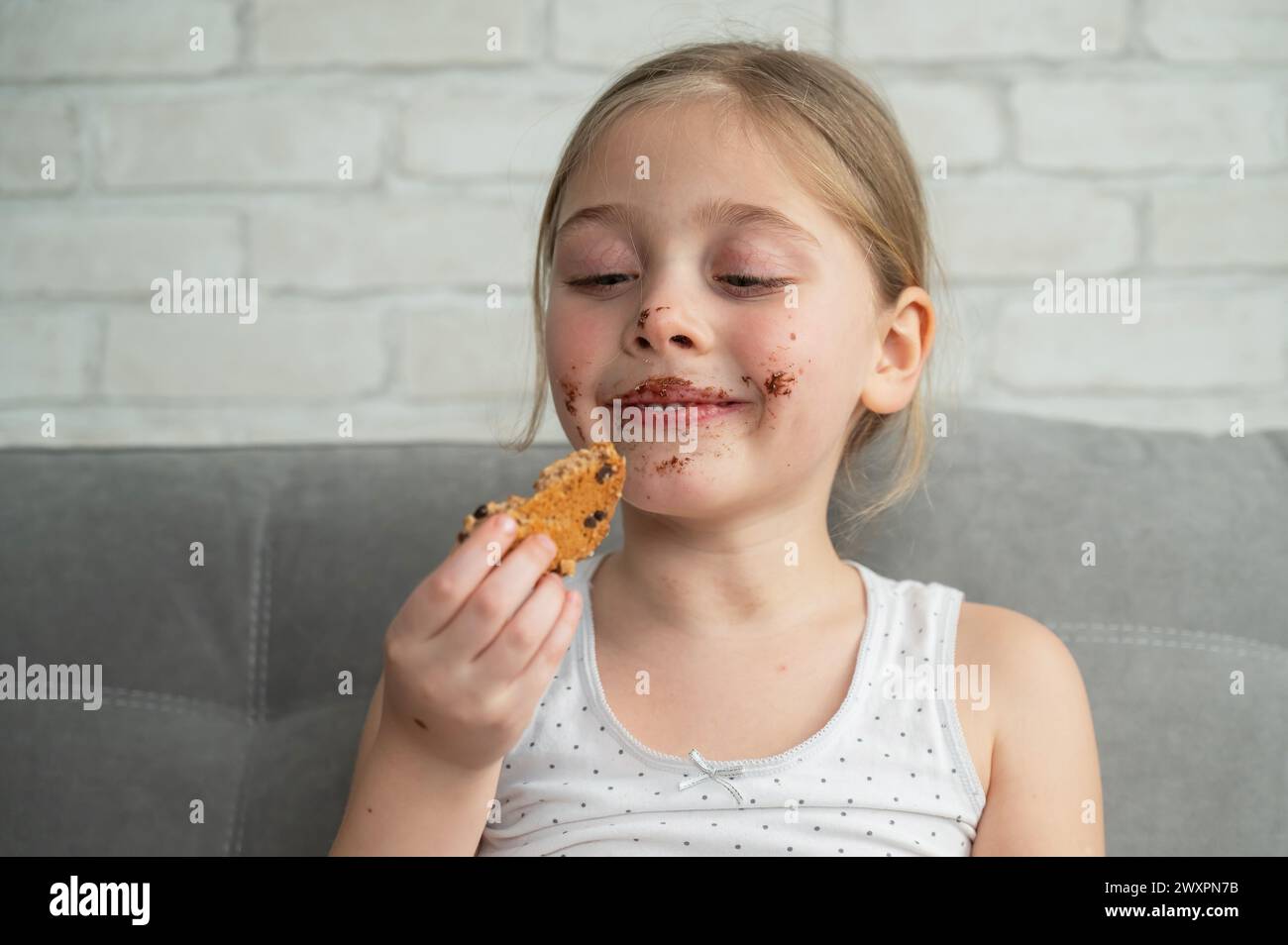 A cute little girl covered in chocolate eats cookies while sitting on the sofa. Stock Photo