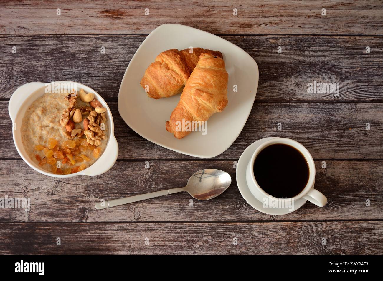 A plate of oatmeal with raisins and nuts, a plate with two fresh croissants and a cup of hot black coffee on a wooden table. Top view, flat lay. Stock Photo