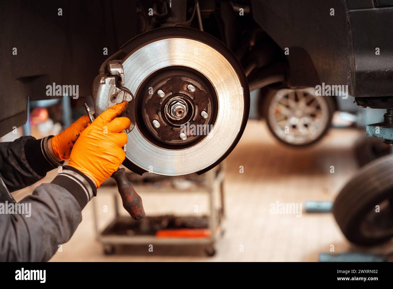 Anonymous mechanic in jacket and gloves while standing in workplace in automobile garage and replacing car wheel brake pad against blurred interior in Stock Photo