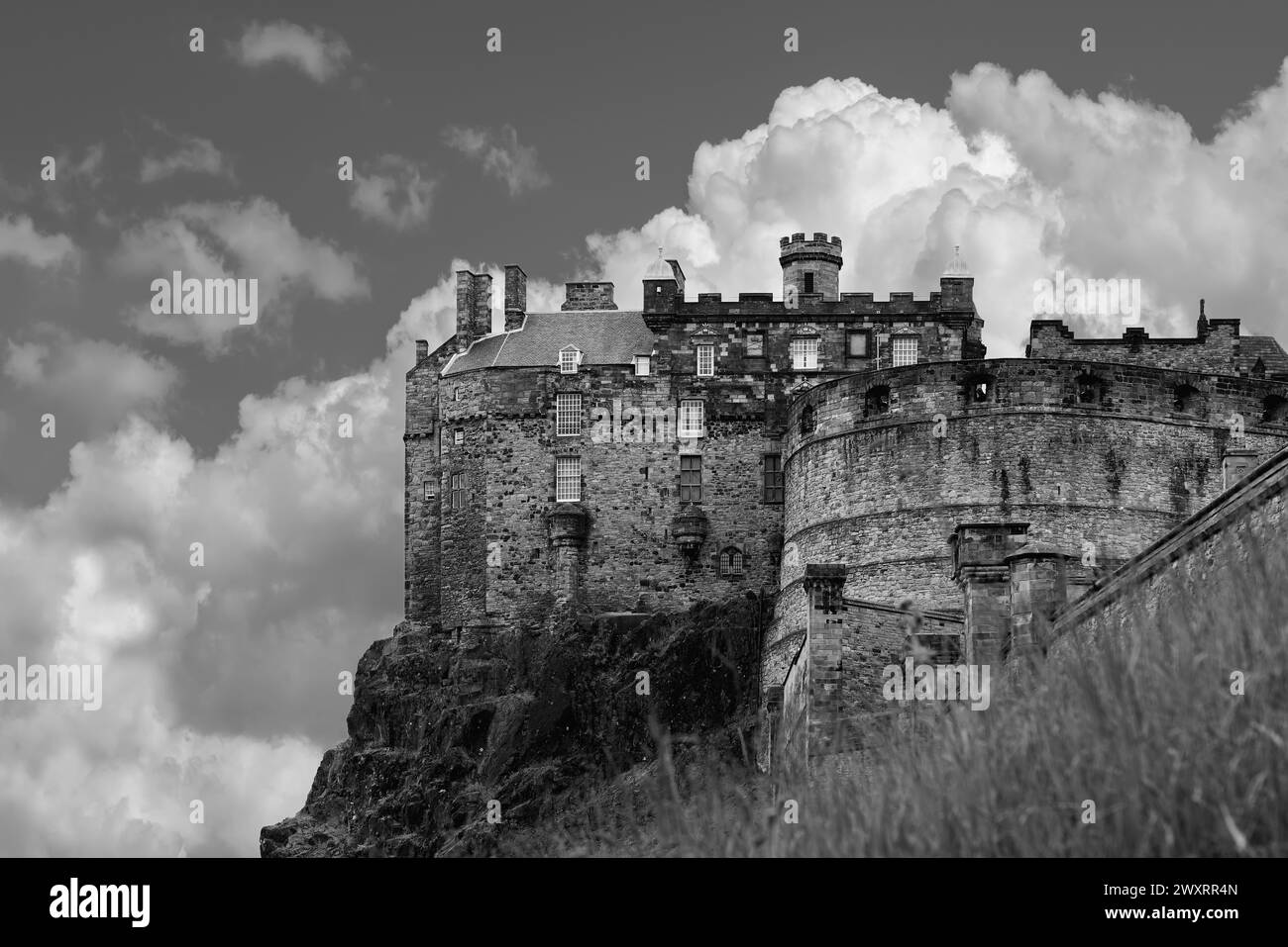 The formidable walls of Edinburgh Castle loom overhead, with cumulus clouds accentuating its commanding presence in this black and white photo Stock Photo