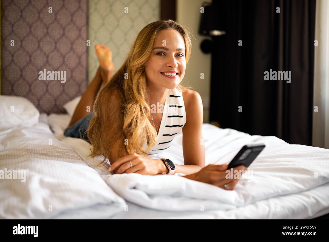 Happy barefoot young woman looking at mobile phone in bed Stock Photo