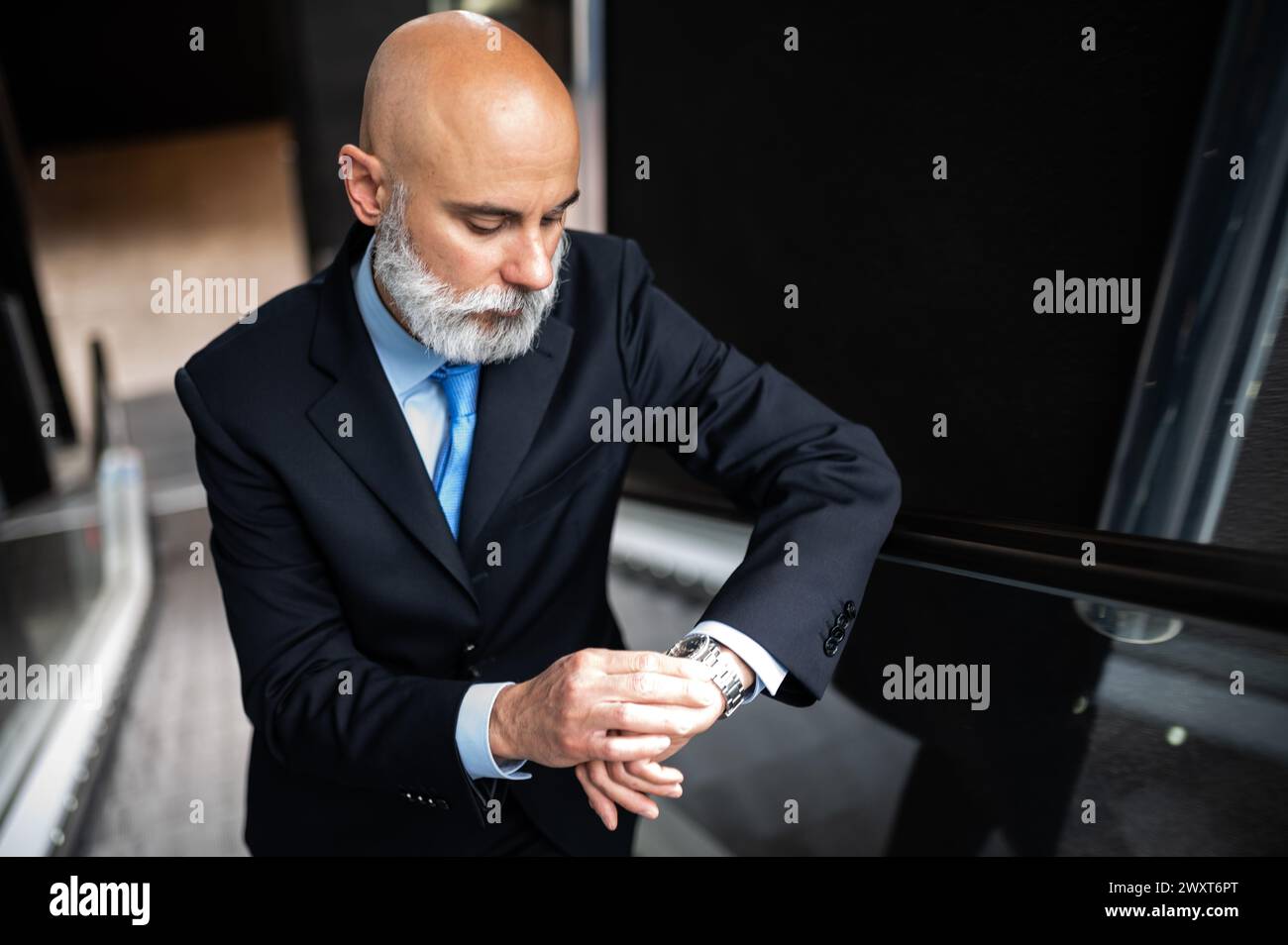 Mature bald stylish business man portrait with a white beard on mobile stairs holding his watch to check time Stock Photo