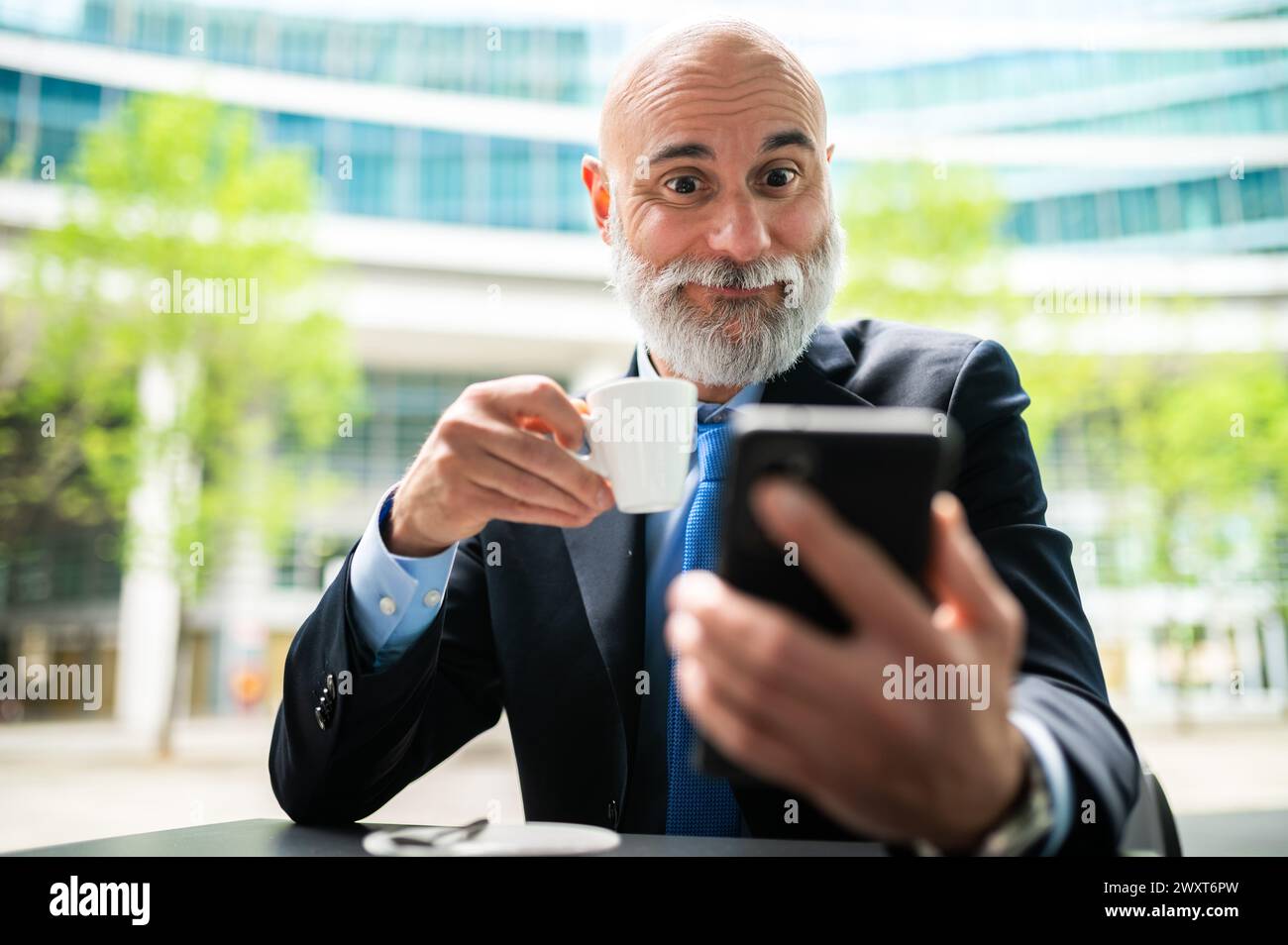Senior stylish bald manager with white beard having a coffee outdoor while using his smartphone with a shocked expression Stock Photo