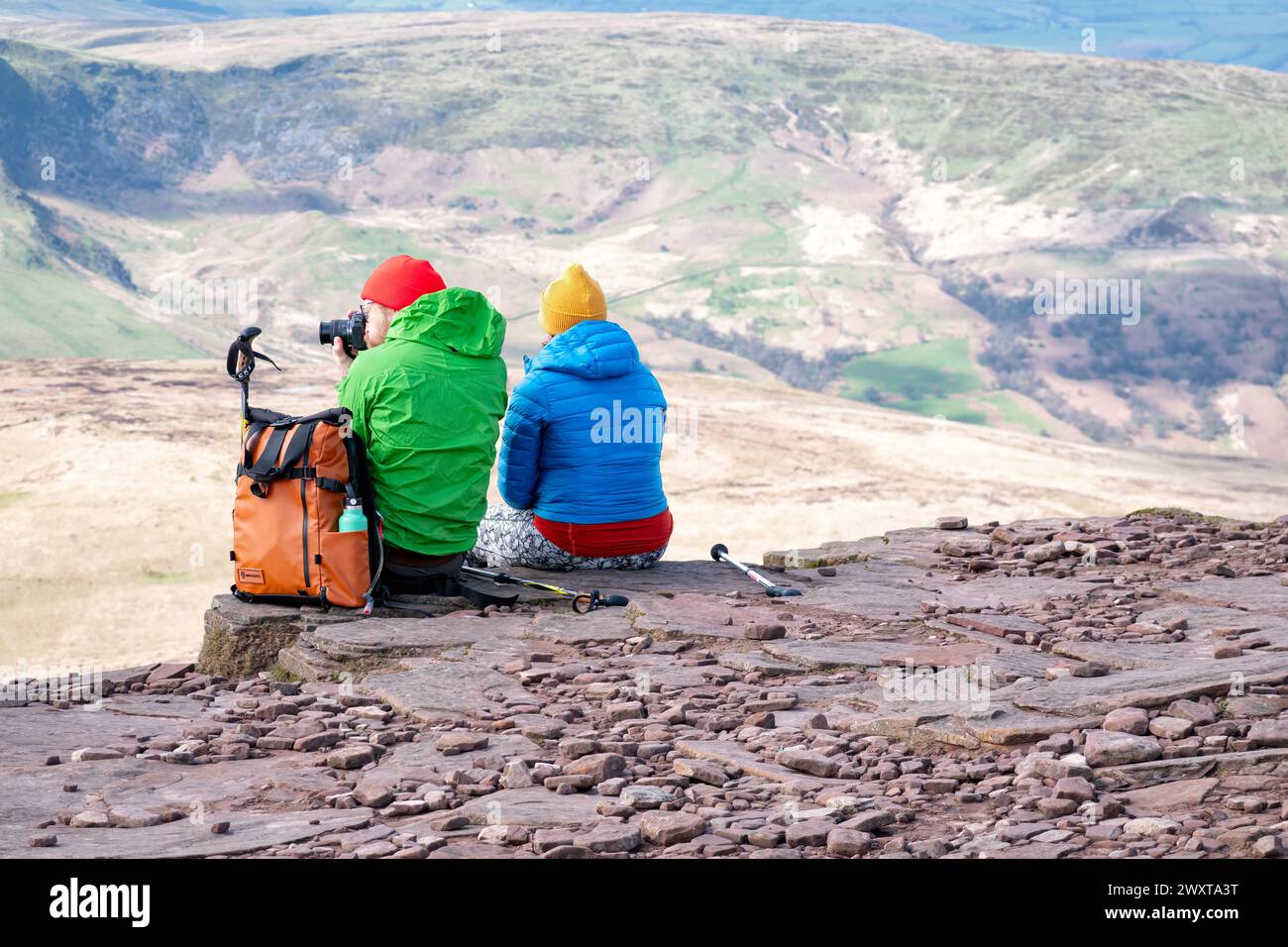 Brecon Beacons, Wales. A male and female walker sit on the ridge of Corn Du at its summit. The man is using a camera to take a picture from the top. Stock Photo