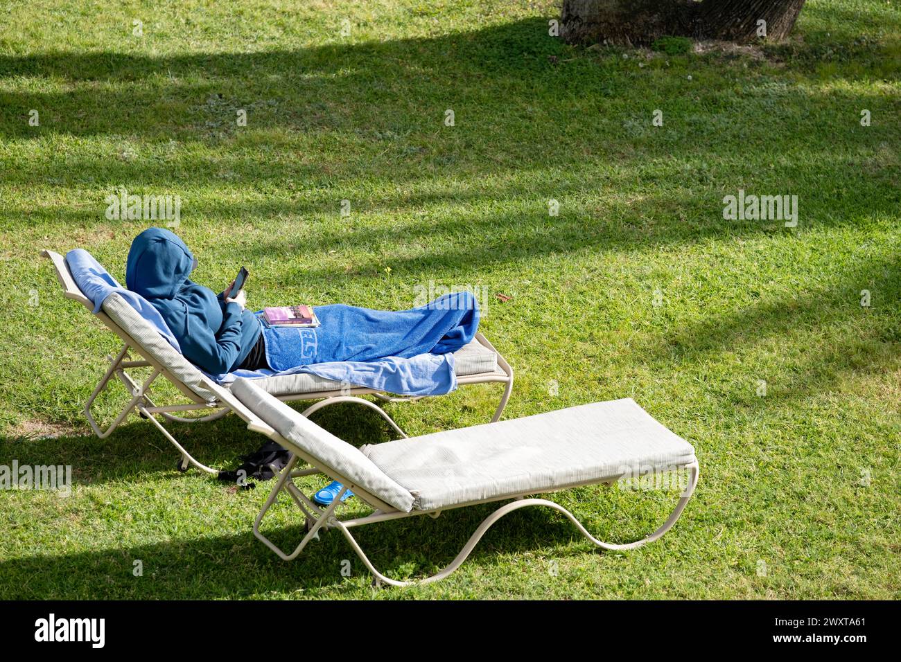 A person lying out on a sun lounger, in sunny weather. The sunbather has wrapped a towel around their legs and is wearing a hooded top to keep warm Stock Photo