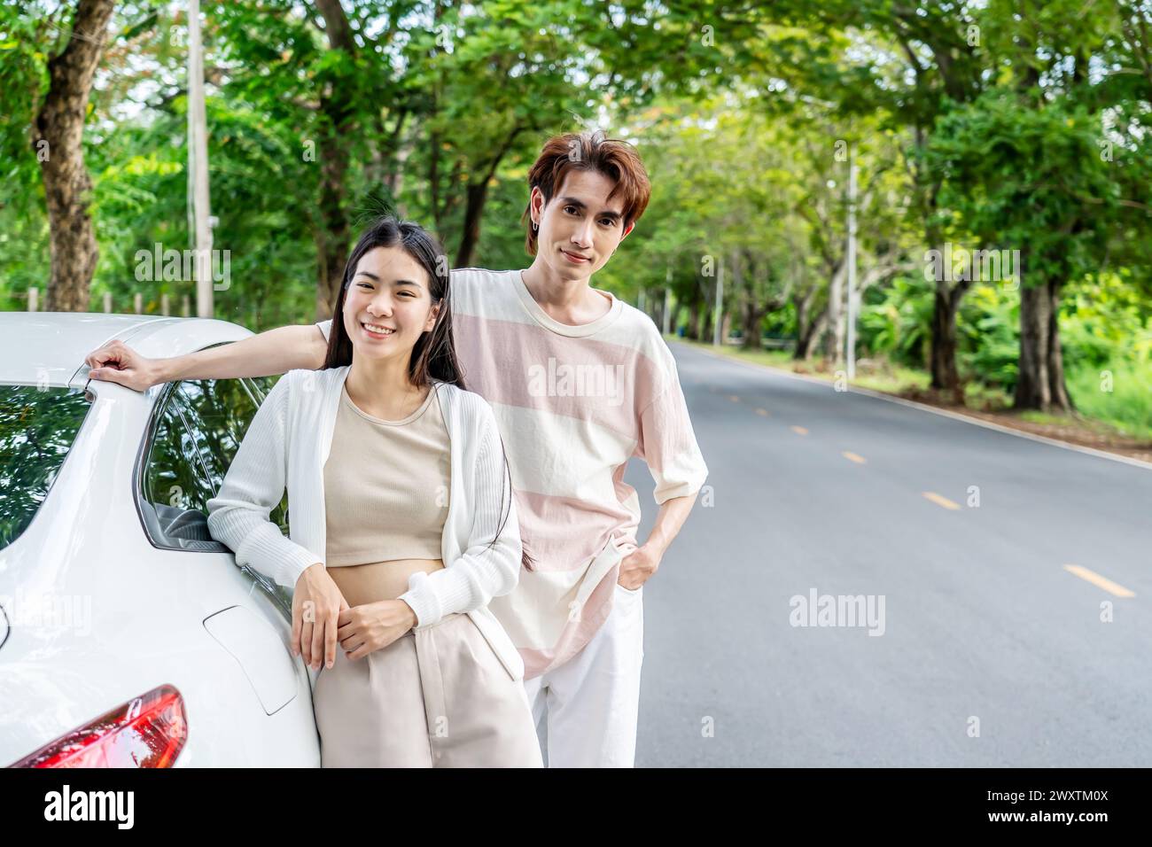 Asian couple standing next to their car on a roadside with lush greenery Stock Photo