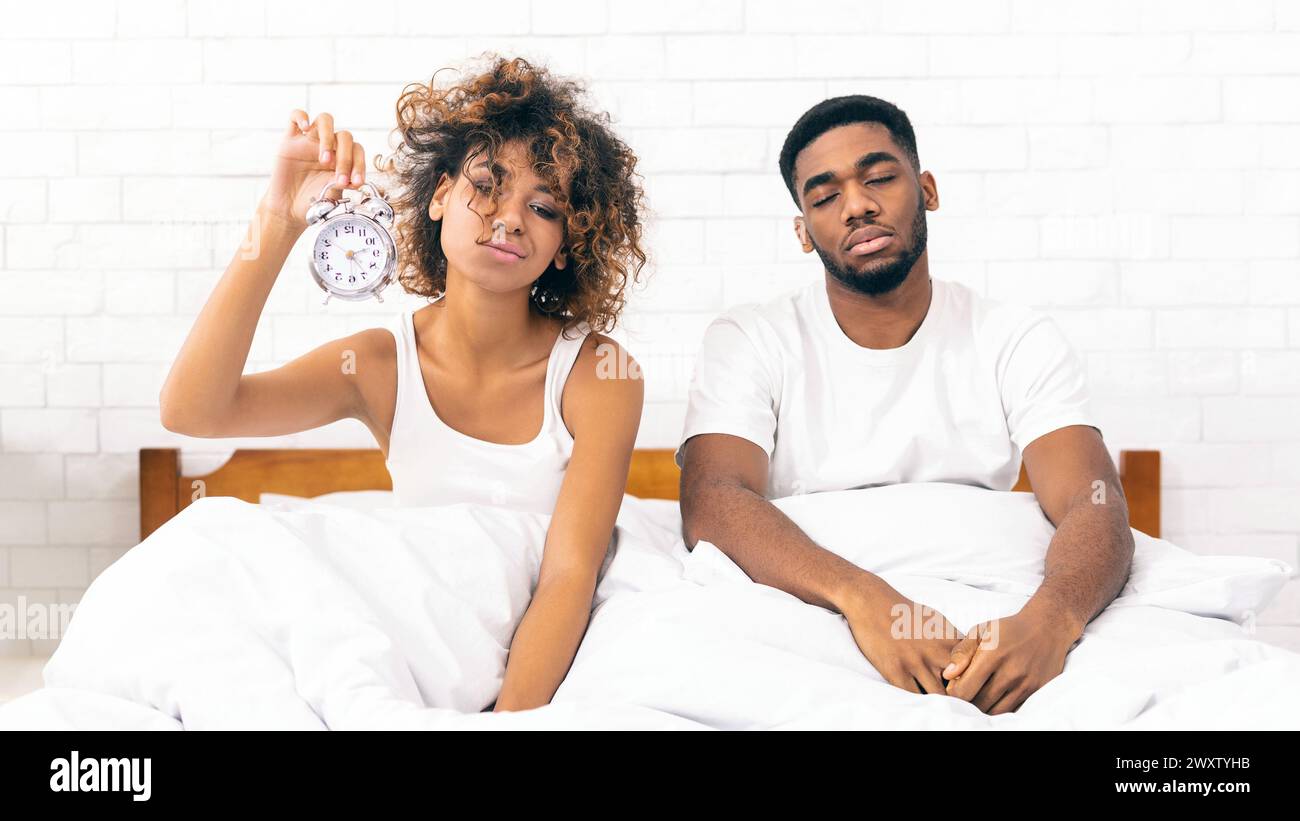 Sleepy african-american couple sitting in bed with clock Stock Photo