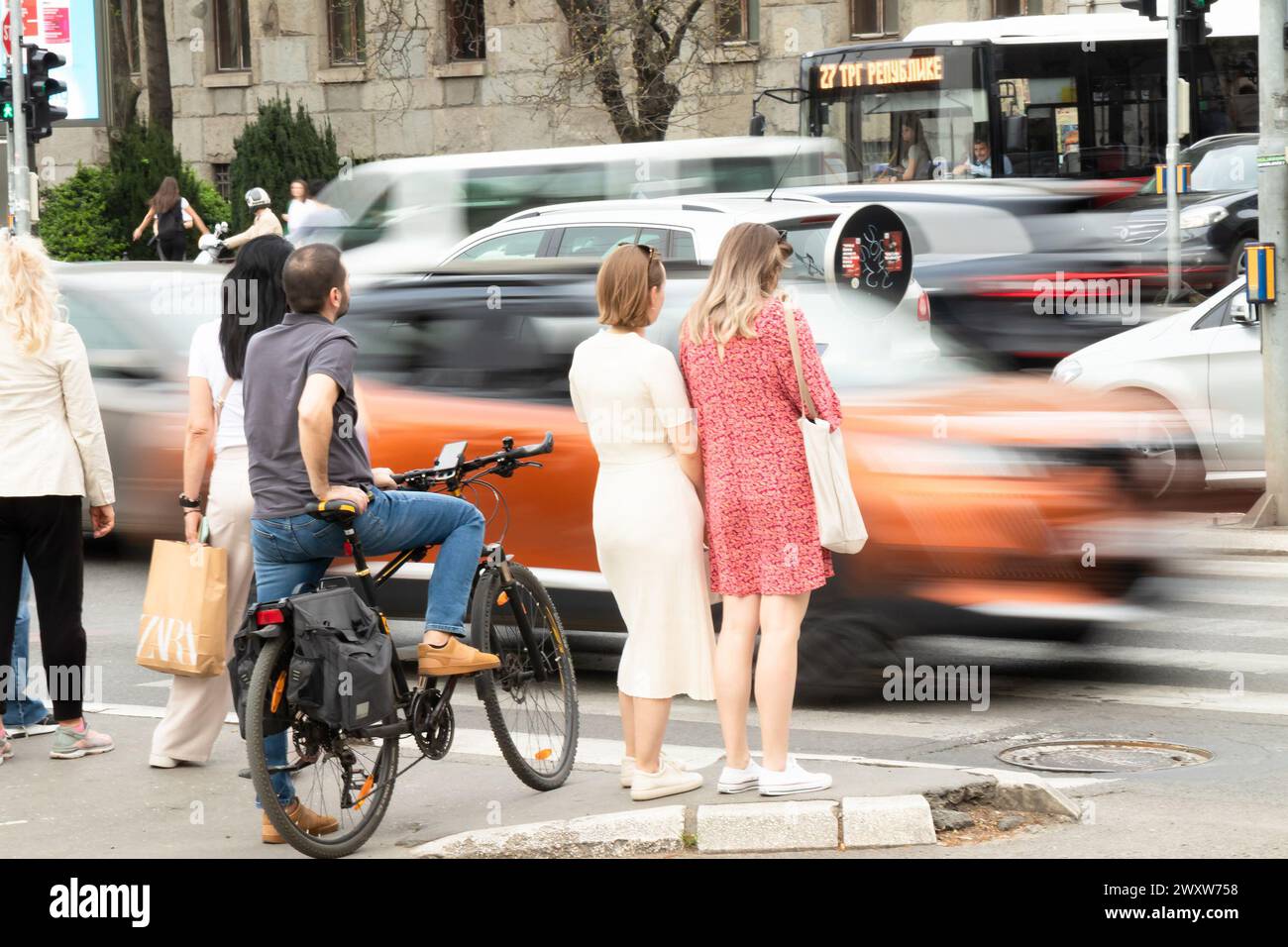 Belgrade, Serbia - April 1, 2024:  People standing on sidewalk, waiting to cross a busy street intersection, rear view with traffic in motion blur on Stock Photo