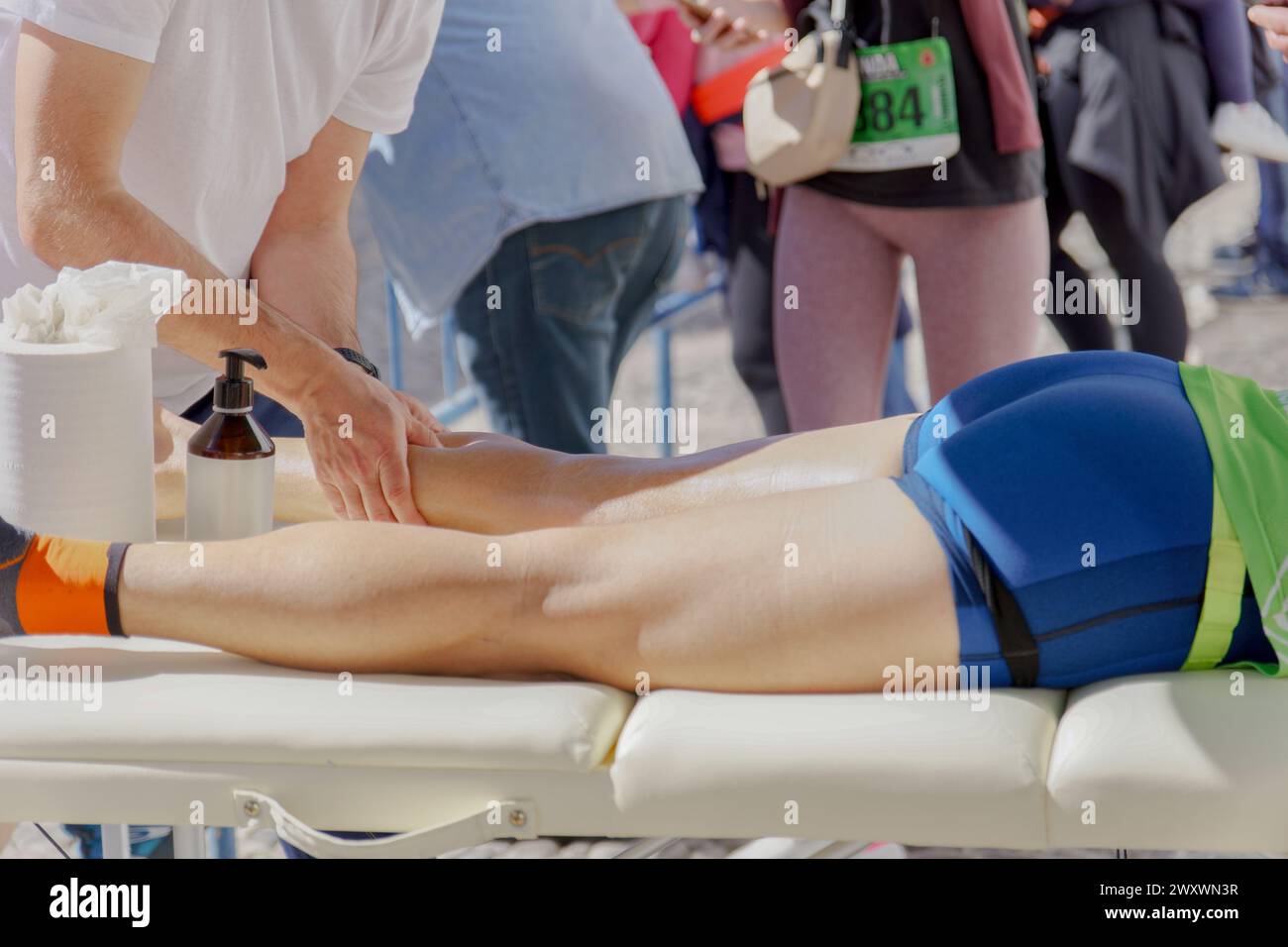 An athletic trainer wearing a white t-shirt is providing a professional leg massage to a runner reclining on a massage table, Stock Photo