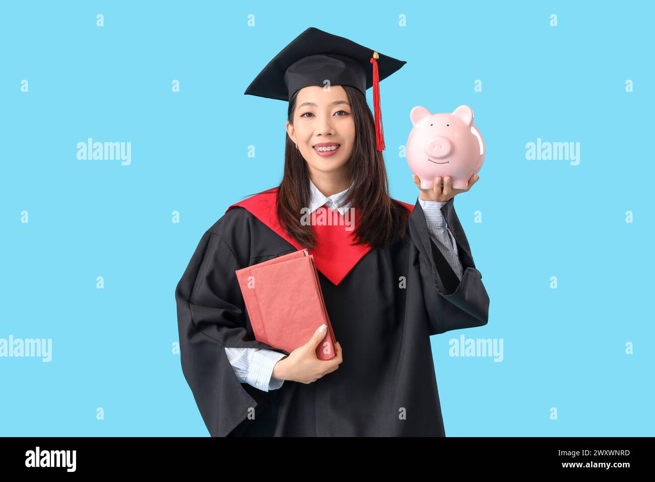 Asian female graduate student with book and piggy bank on blue background. Tuition fees concept Stock Photo