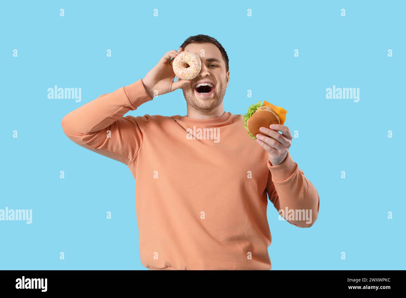 Young man with doughnut and burger on blue background. Overeating concept Stock Photo