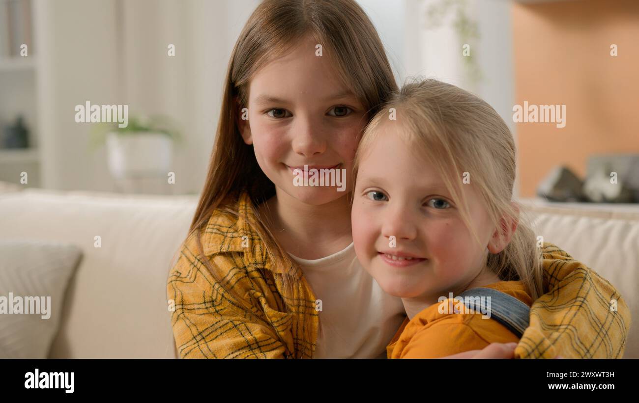 Two cute smiling caucasian kids little girls children sisters friends hugging embracing cuddle embrace hug at home on couch looking camera posing in Stock Photo