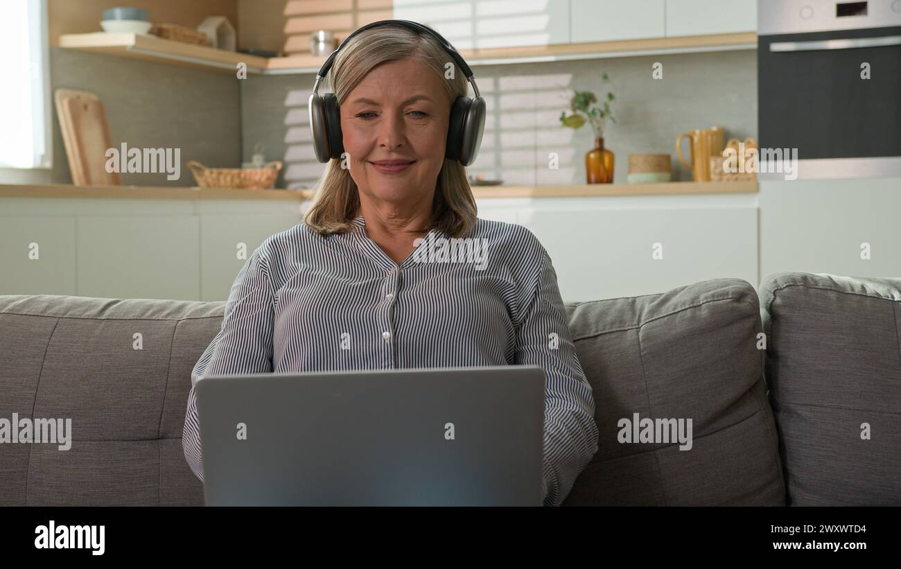 Smiling mature Caucasian woman grandmother businesswoman freelancer typing on laptop at home listening music on headphones communicate on social Stock Photo
