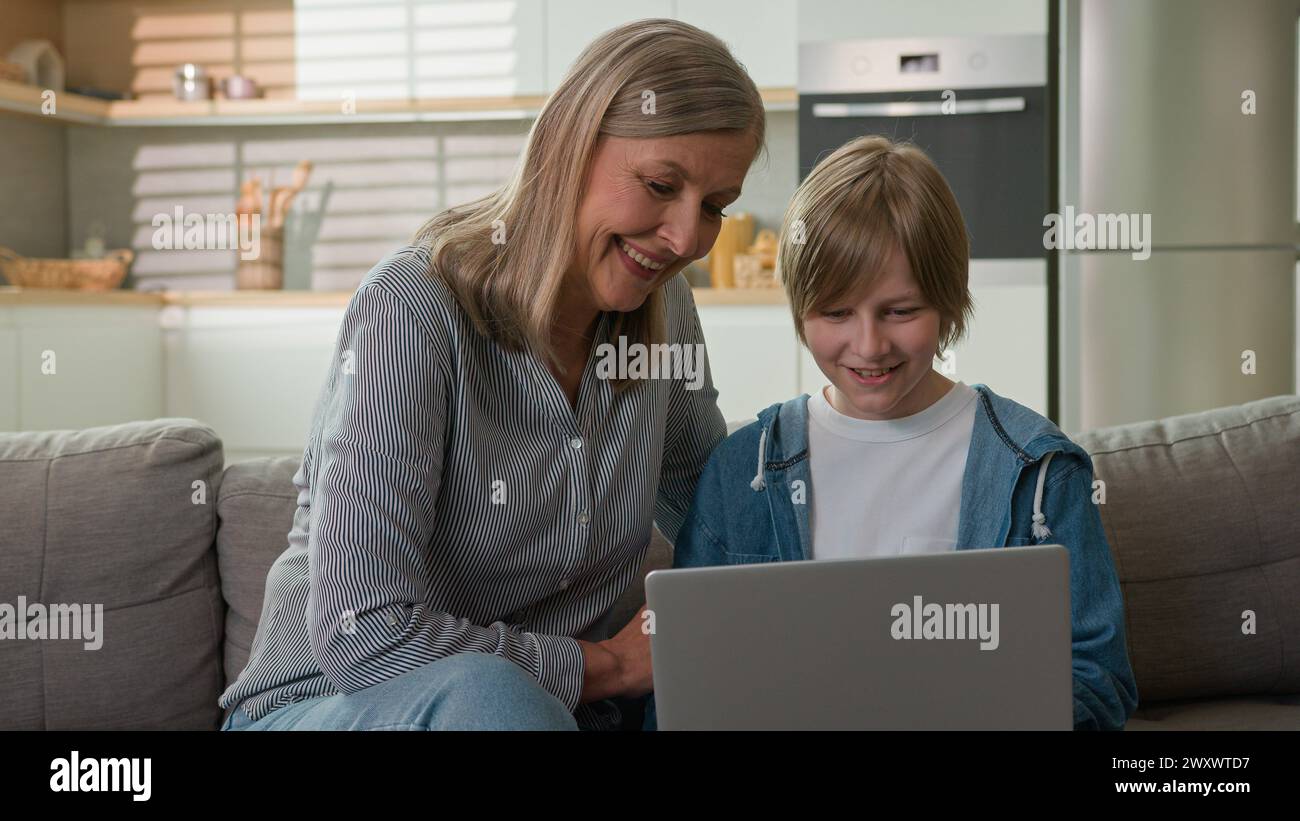 Caucasian elderly woman 50s with grandson browsing social media on laptop shopping in online store talking smiling laughing at home couch caring Stock Photo