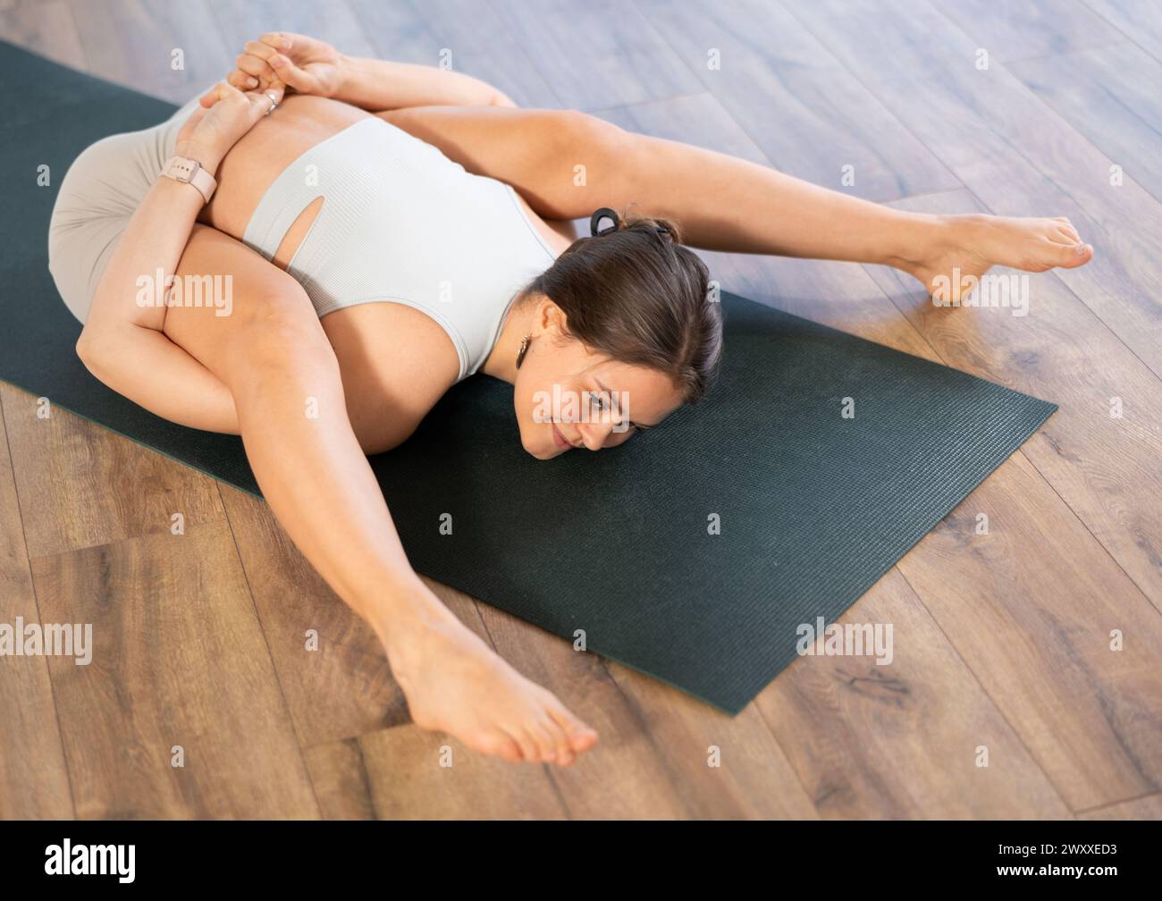 Happy young woman doing difficult pose of yoga, Tortoise Pose on mat in yoga studio Stock Photo