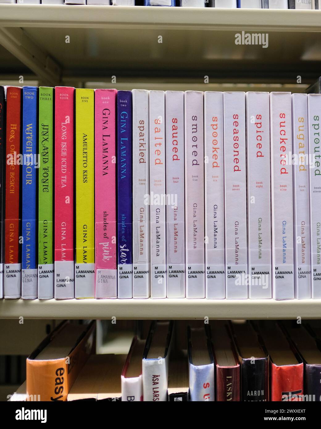 Library bookshelf with murder mystery books by American author Gina LaManna; the Lacey Luzzi Mafia Mystery and Magic & Mixology Mystery series. Stock Photo