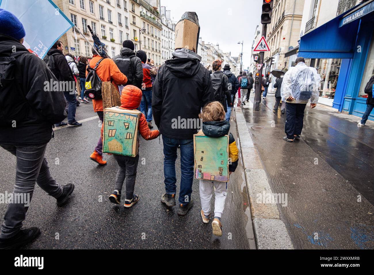 A man with a paper house on his head walks with his two kids with paper houses inform of backpacks, during the demonstration against the housing crisis. Thousands of people attended the demonstration for the right to housing, in Paris. The movement organized by the association 'DAL' Droit Au Logement (Right to Housing), focused on denouncing the start of evictions that will begin at the beginning of April. It was also demanded a reduction in rents, the requisition of empty houses for rent, accommodation for all inhabitants and an end to real estate speculation. Stock Photo