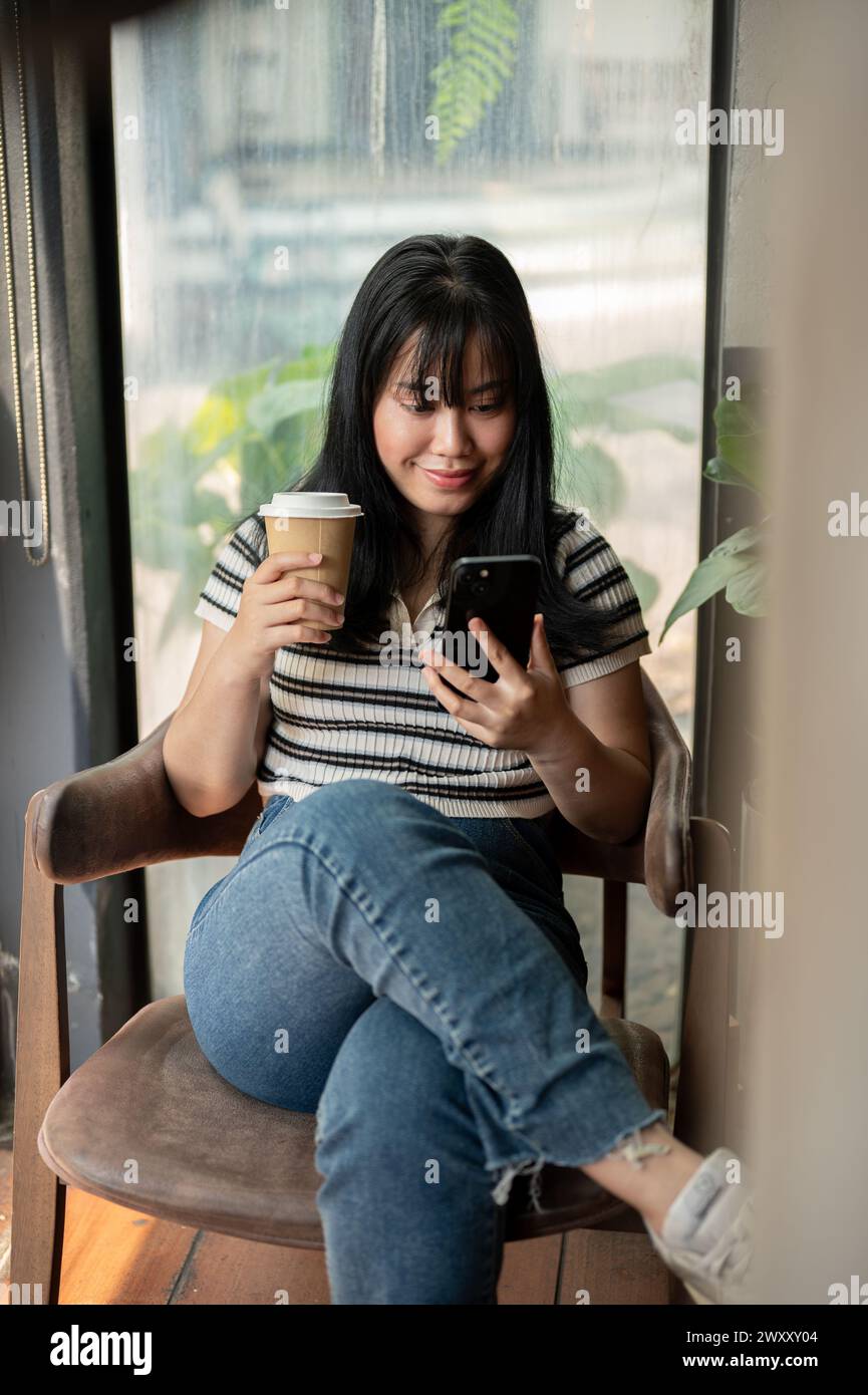 A positive young Asian woman relaxing in a cafe, using her smartphone ...