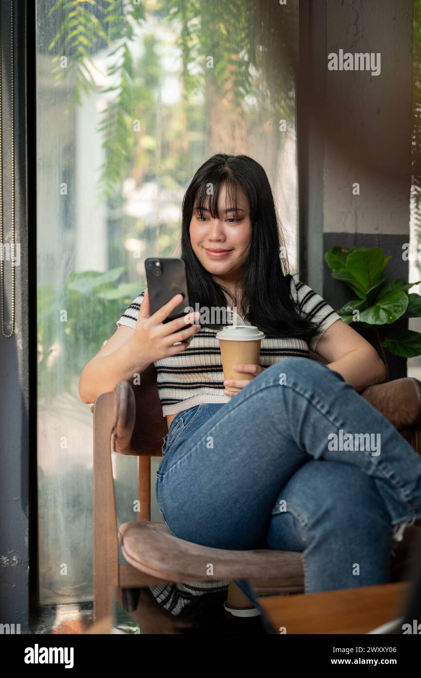 A positive young Asian woman relaxing in a cafe, using her smartphone ...