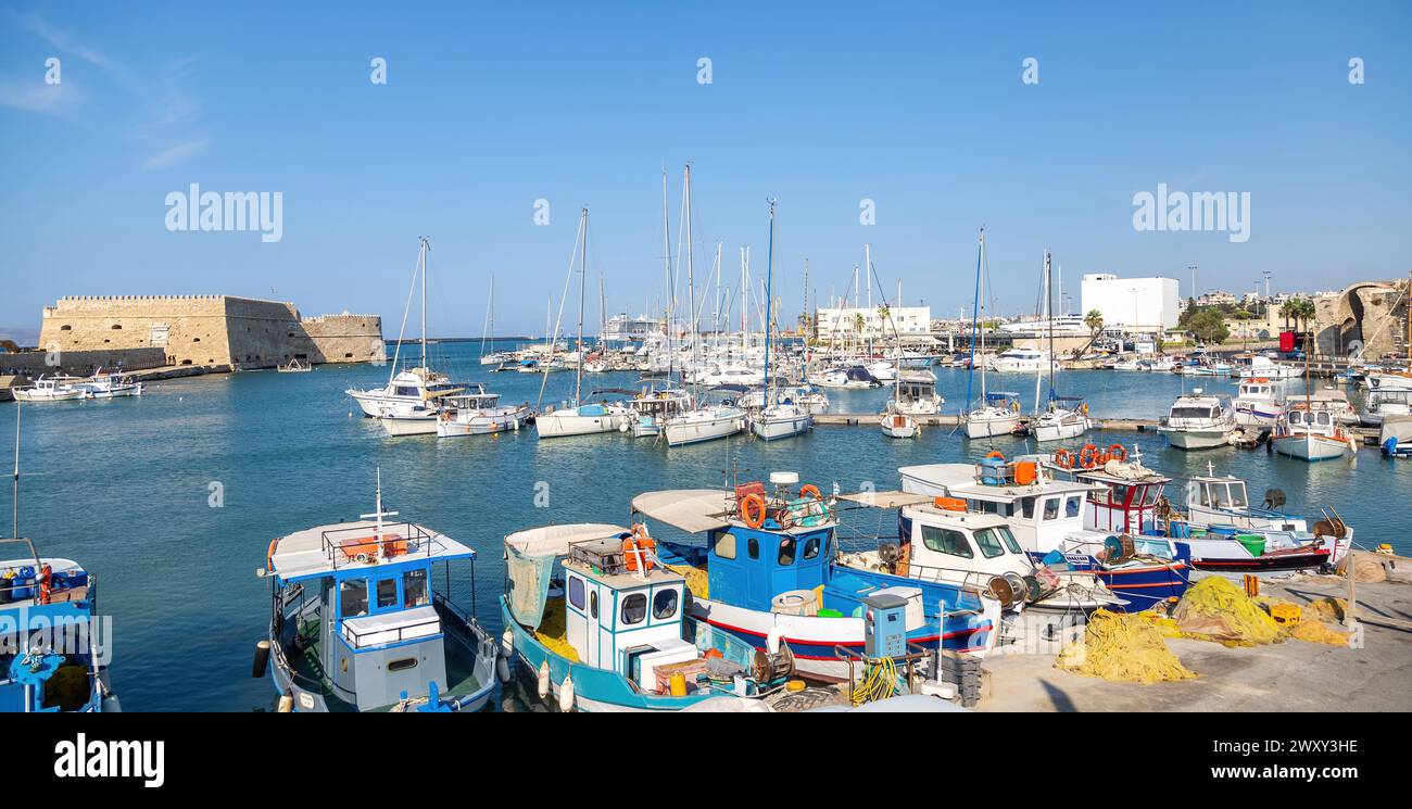 Crete island, destination Greece. Moored ship and boat with mast at Heraklion old Venetian harbor, calm sea water, blue sky background. Stock Photo