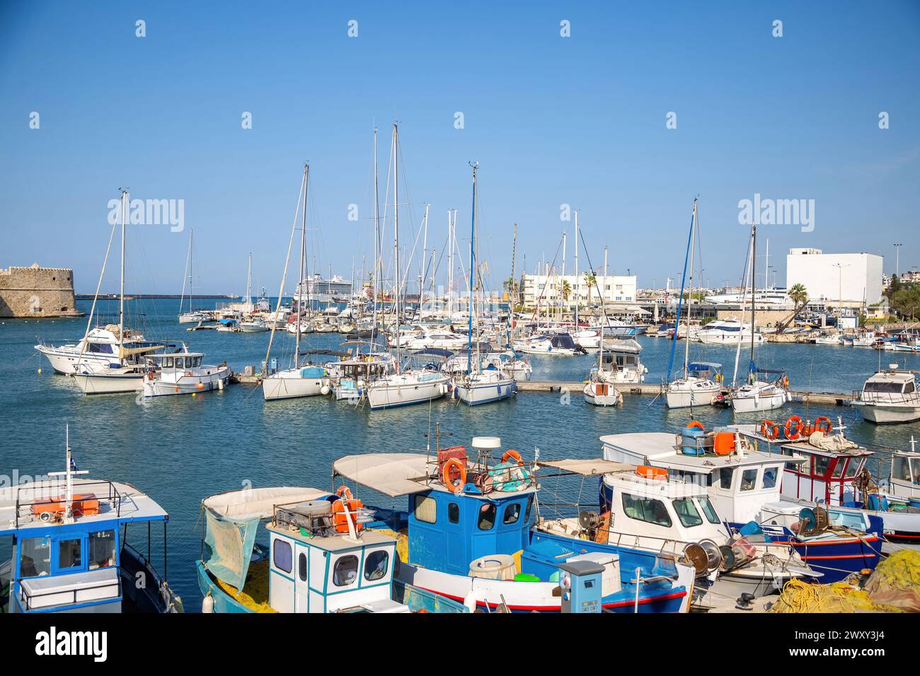 Crete island, destination Greece. Moored vessel with mast and fishing boat at Heraklion old Venetian harbor, calm sea water, blue sky background. Stock Photo