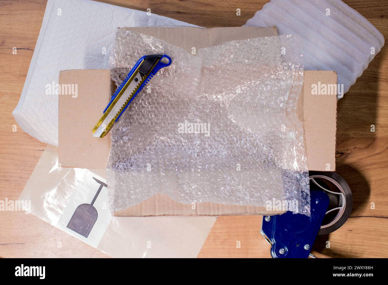 Open box with wrapped items, adhesive tape, scissors, paper and bubble wrap on wooden table, flat lay. Stock Photo
