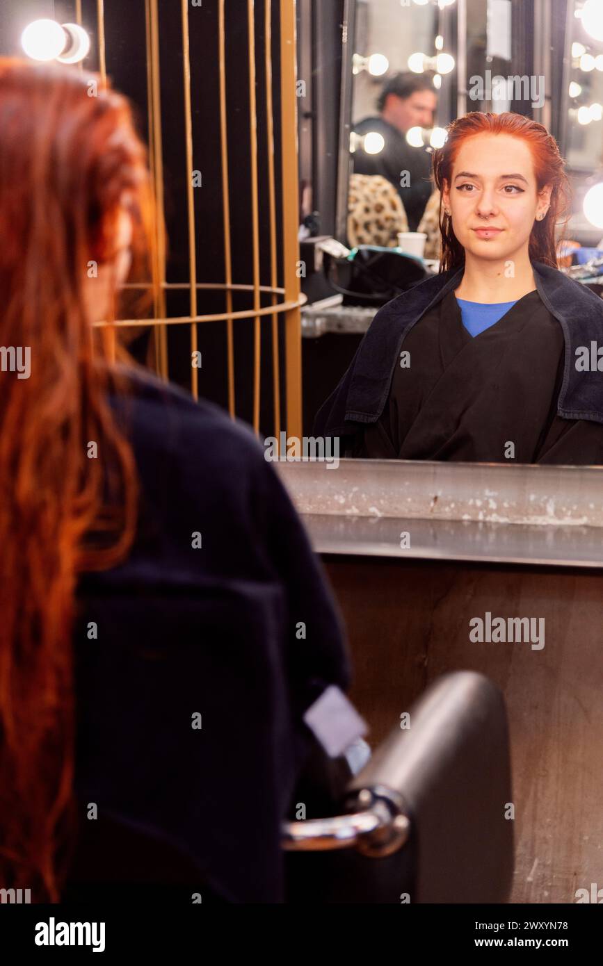 A young client with red hair sits in front of a salon mirror, ready for a hair styling session, reflecting the salon's vibrant atmosphere Stock Photo