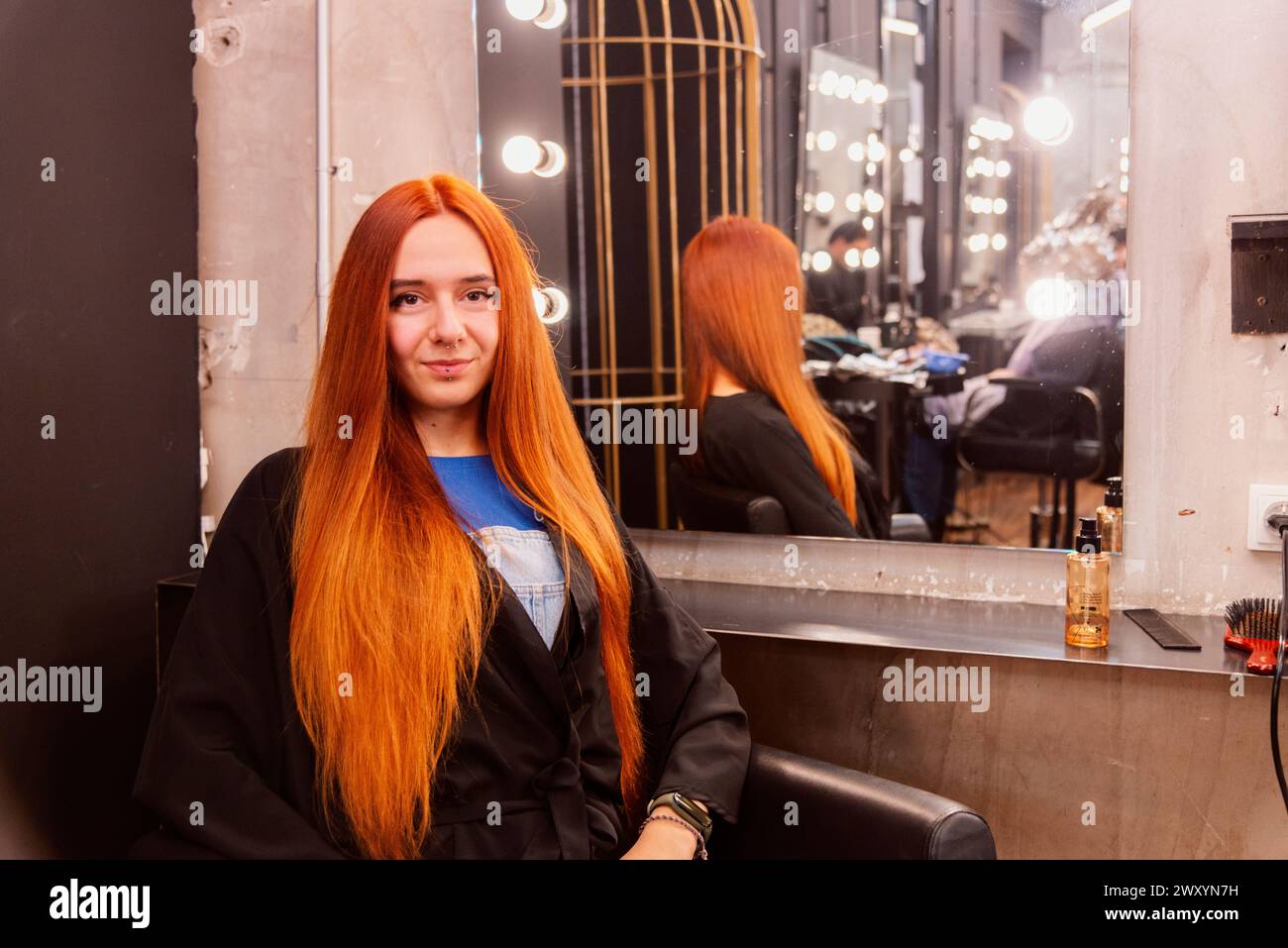 A young client with vibrant red hair sits in a chic hair salon, smiling gently, reflecting on a mirror Stock Photo