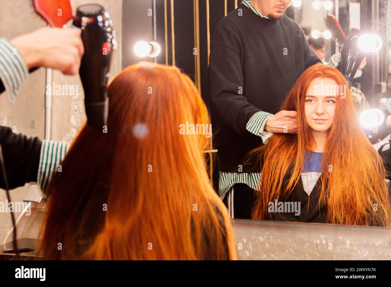 A hairstylist is drying a young woman's long, red hair with a blow dryer at a professional hair salon, as the client watches the process in a mirror Stock Photo