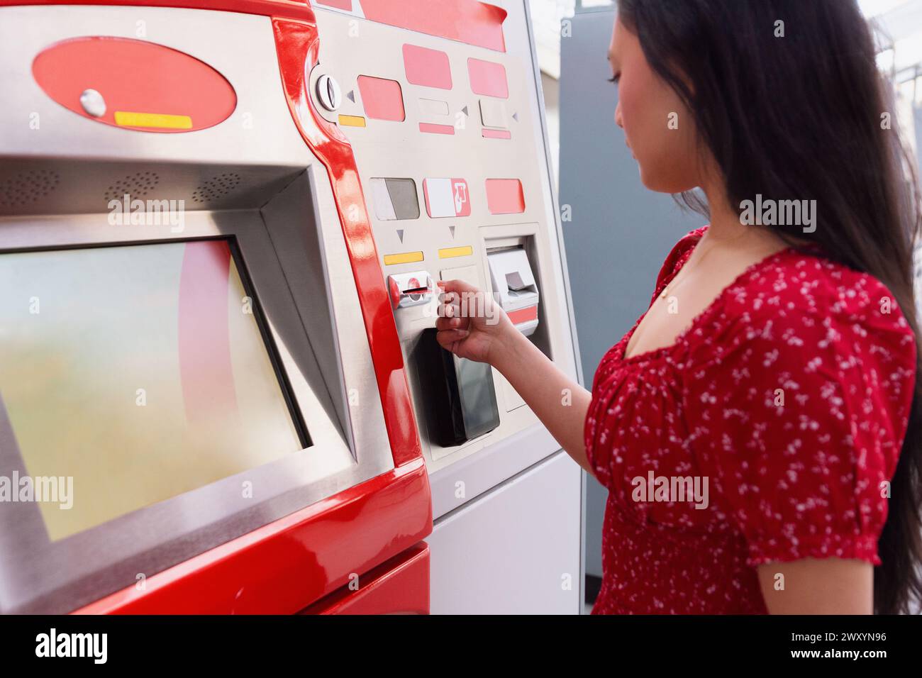 A woman in a vibrant red dress is purchasing a ticket from a modern, red vending machine at a station Stock Photo