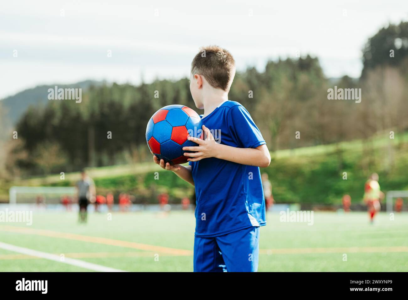 A young boy in a blue jersey lovingly holds a soccer ball, watching a match on a sunny field Stock Photo