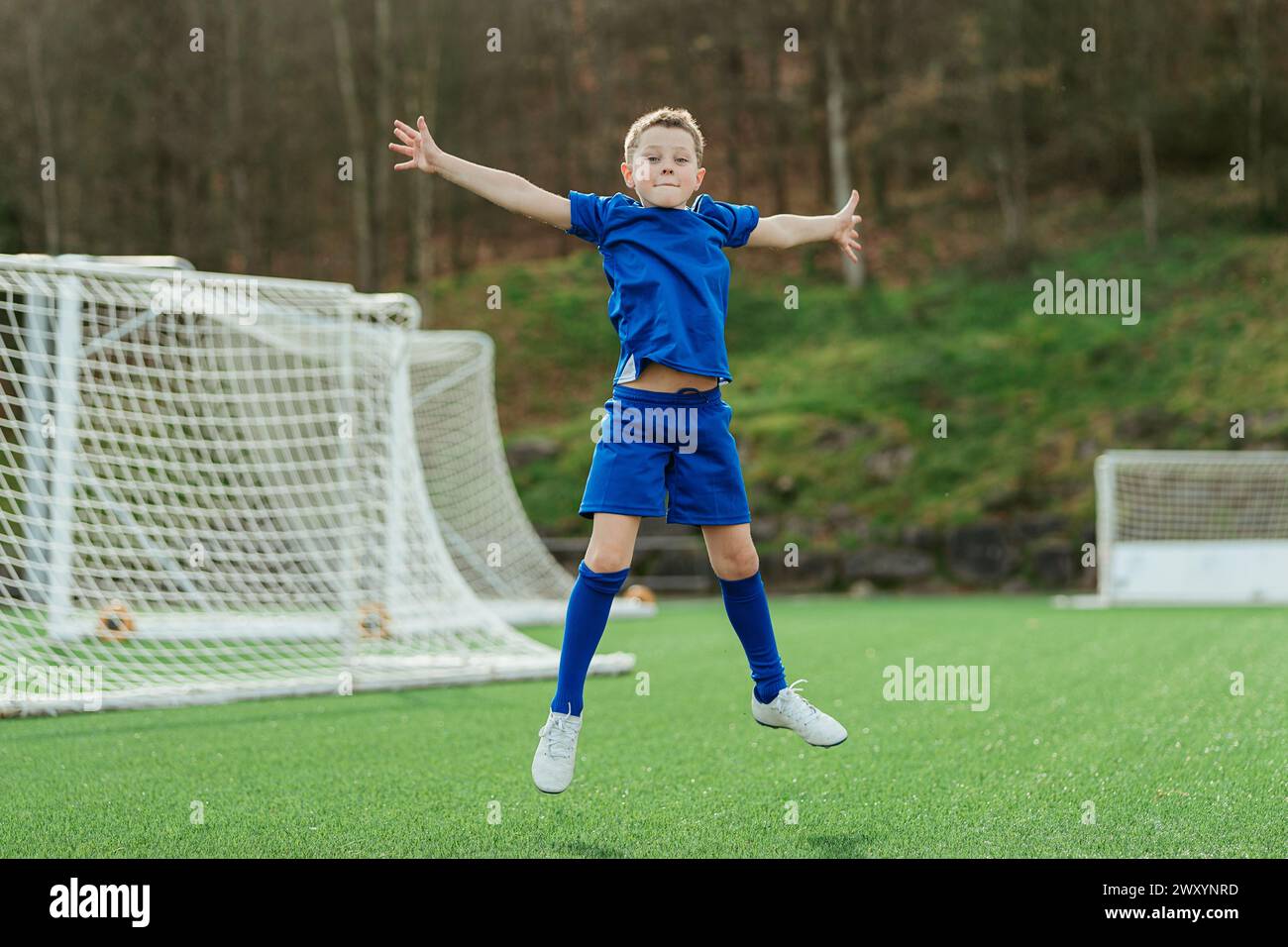 A young boy in a blue soccer uniform joyfully celebrates with arms outstretched on a soccer field, with a goal net in the background Stock Photo