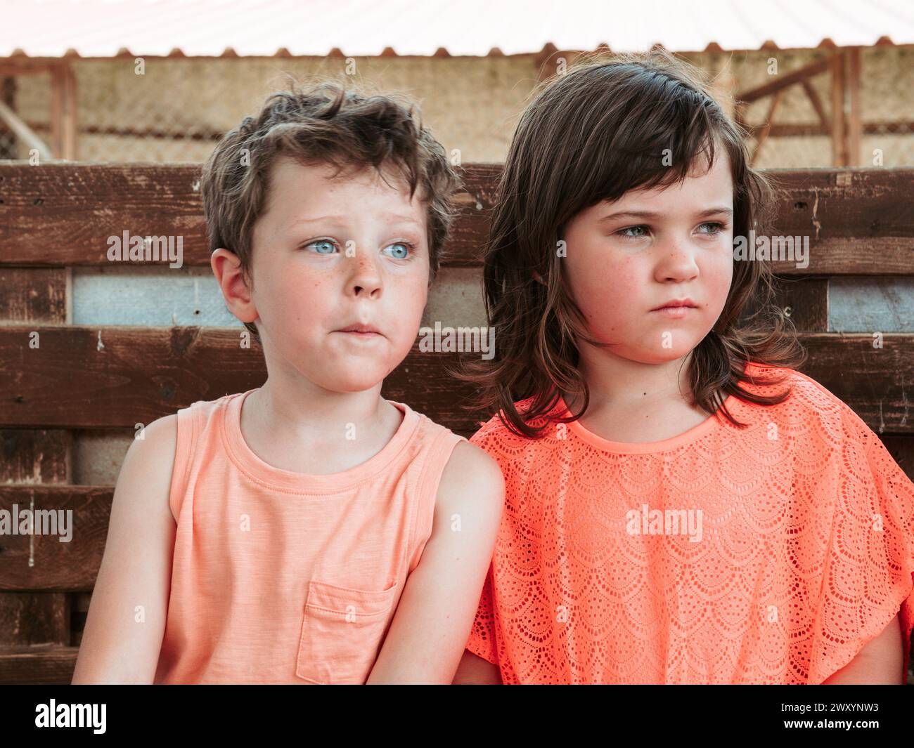 A boy and girl sit side by side against a wooden fence, lost in thought and taking in the sights of a farm school during summer Stock Photo