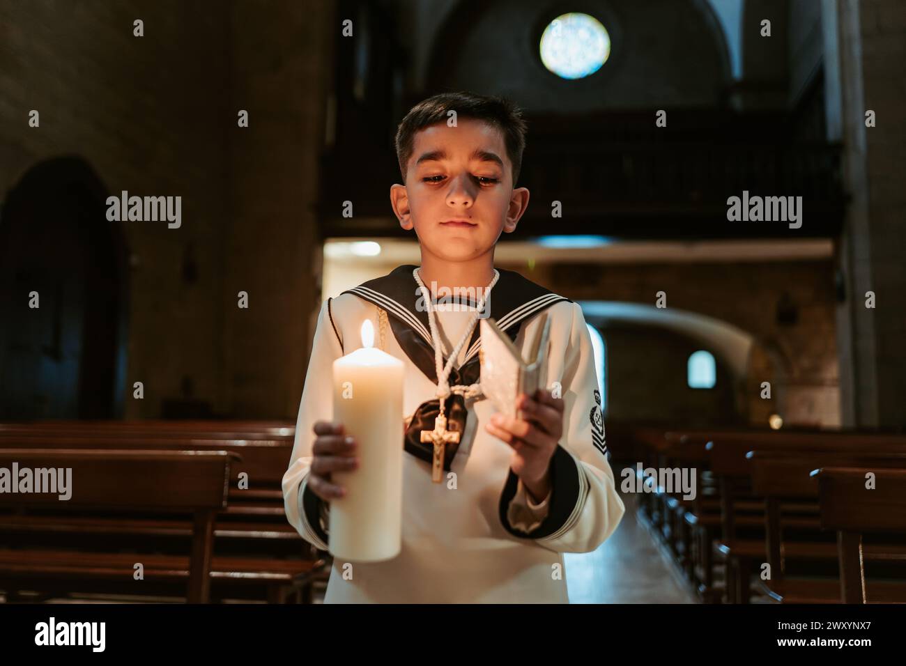 A solemn young boy, dressed in traditional First Communion garb, holds ...