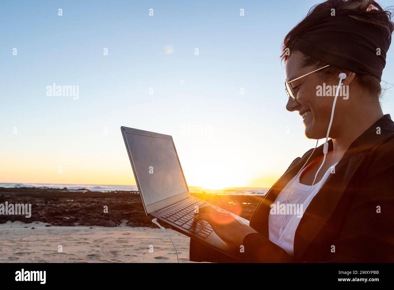 Side view of smiling woman works on her laptop on a beach at sunset, embodying the digital nomad lifestyle with headphones on and the glow of the sett Stock Photo