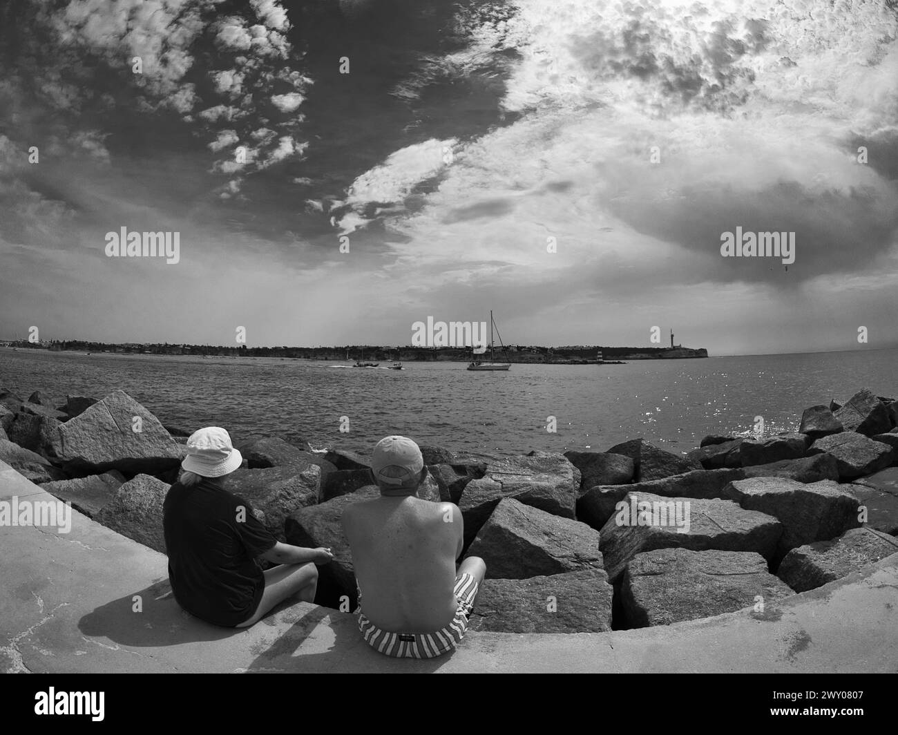 A couple of tourists sitting on the ground looking at the sea and river with boats passing by. Stock Photo