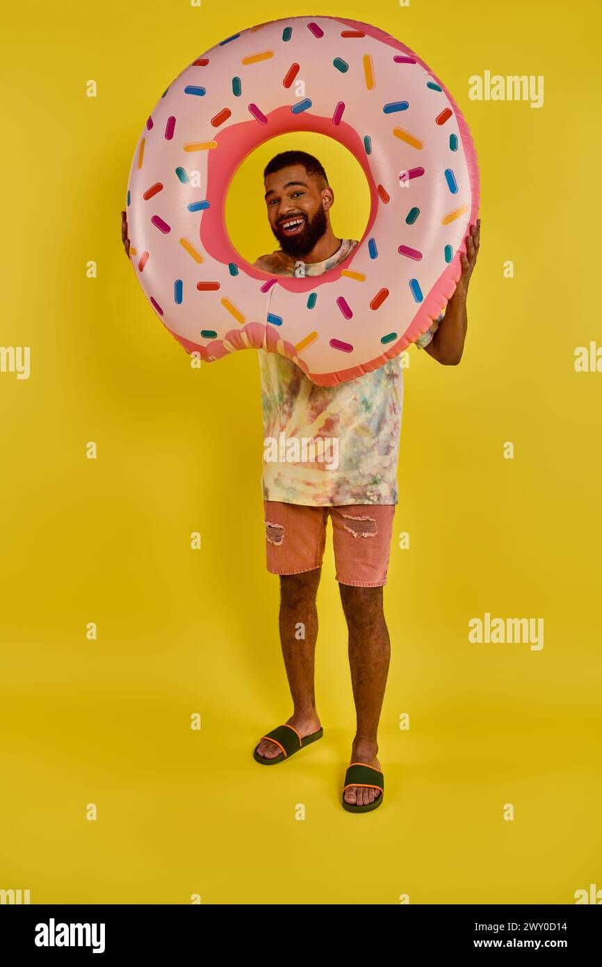 A man playfully holds a giant donut in front of his face, covering it completely. The colorful sprinkles contrast with his expression of joy. Stock Photo