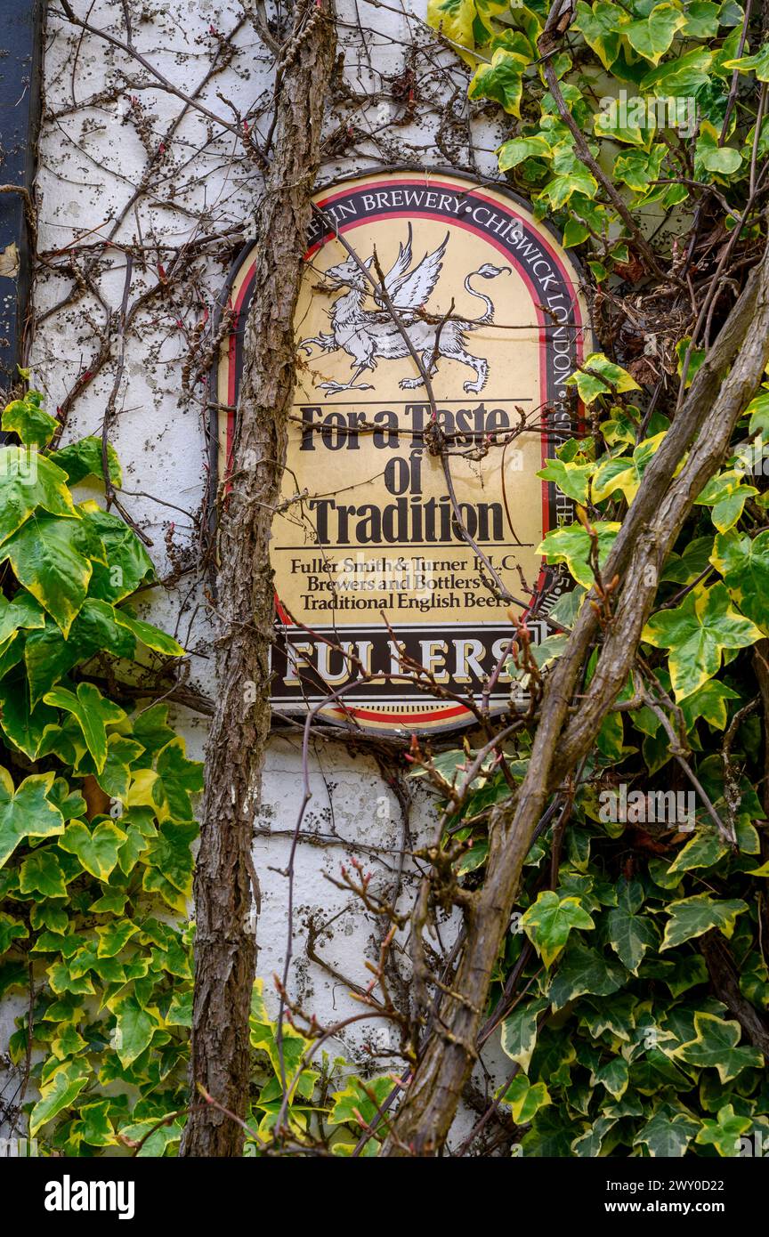 Vintage enamel sign for Fullers beers partly covered by vines on the wall of The Rising Sun pub in Nutbourne village in West Sussex, England. Stock Photo