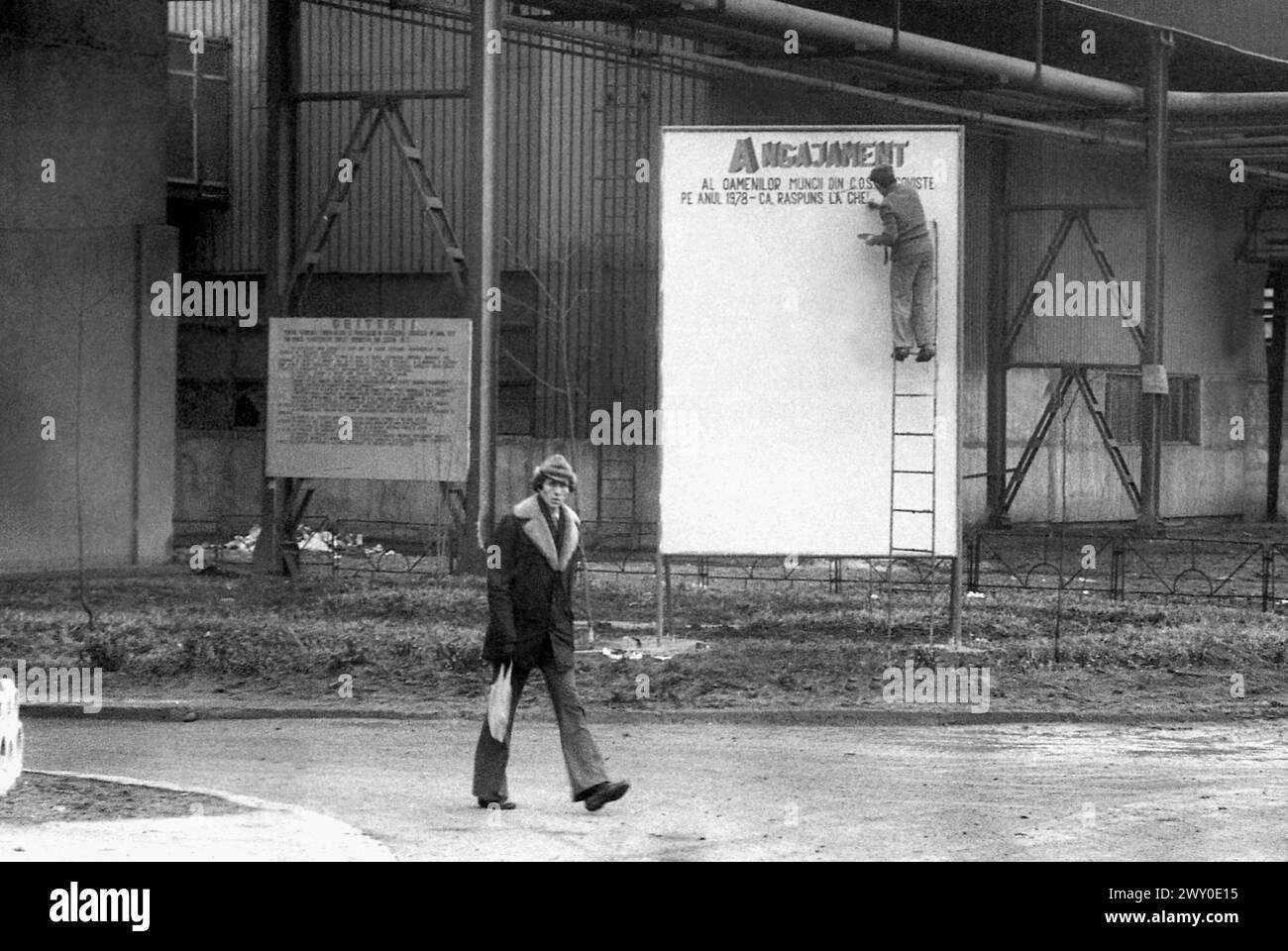 Socialist Republic of Romania in the 1970s. A worker is walking on the grounds of a state-owned factory, while behind him another worker is handwriting on a large board a communist propagandistic message that starts with the words 'Commitment of the working people...'. Stock Photo