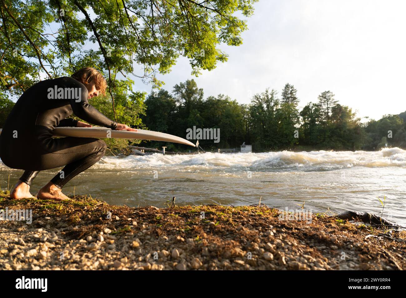 A young surfer kneels by the riverbank with his surfboard, waves crashing during the Aare River flood. Stock Photo
