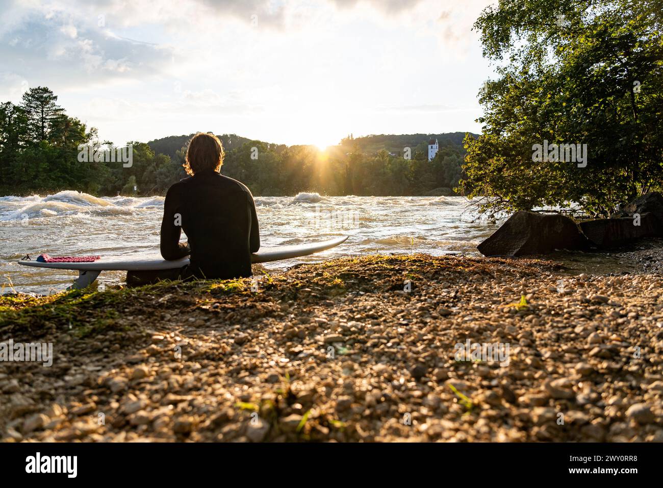 A young surfer sits by the Aare River, gazing at the sunset, enjoying the calm ambiance. Stock Photo