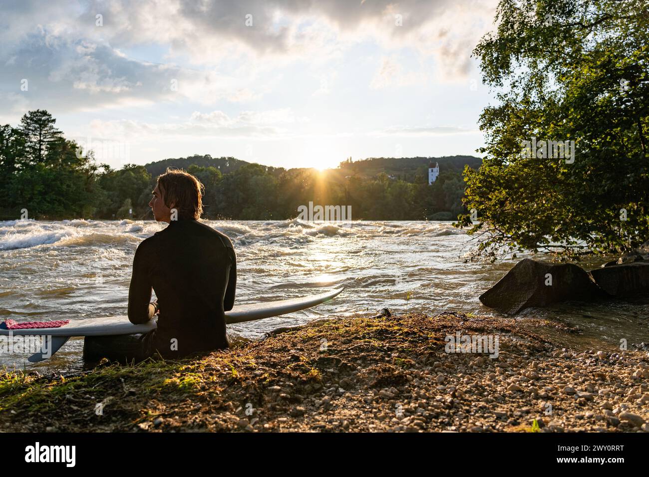A young surfer sits by the Aare River, gazing at the sunset, enjoying the calm ambiance. Stock Photo