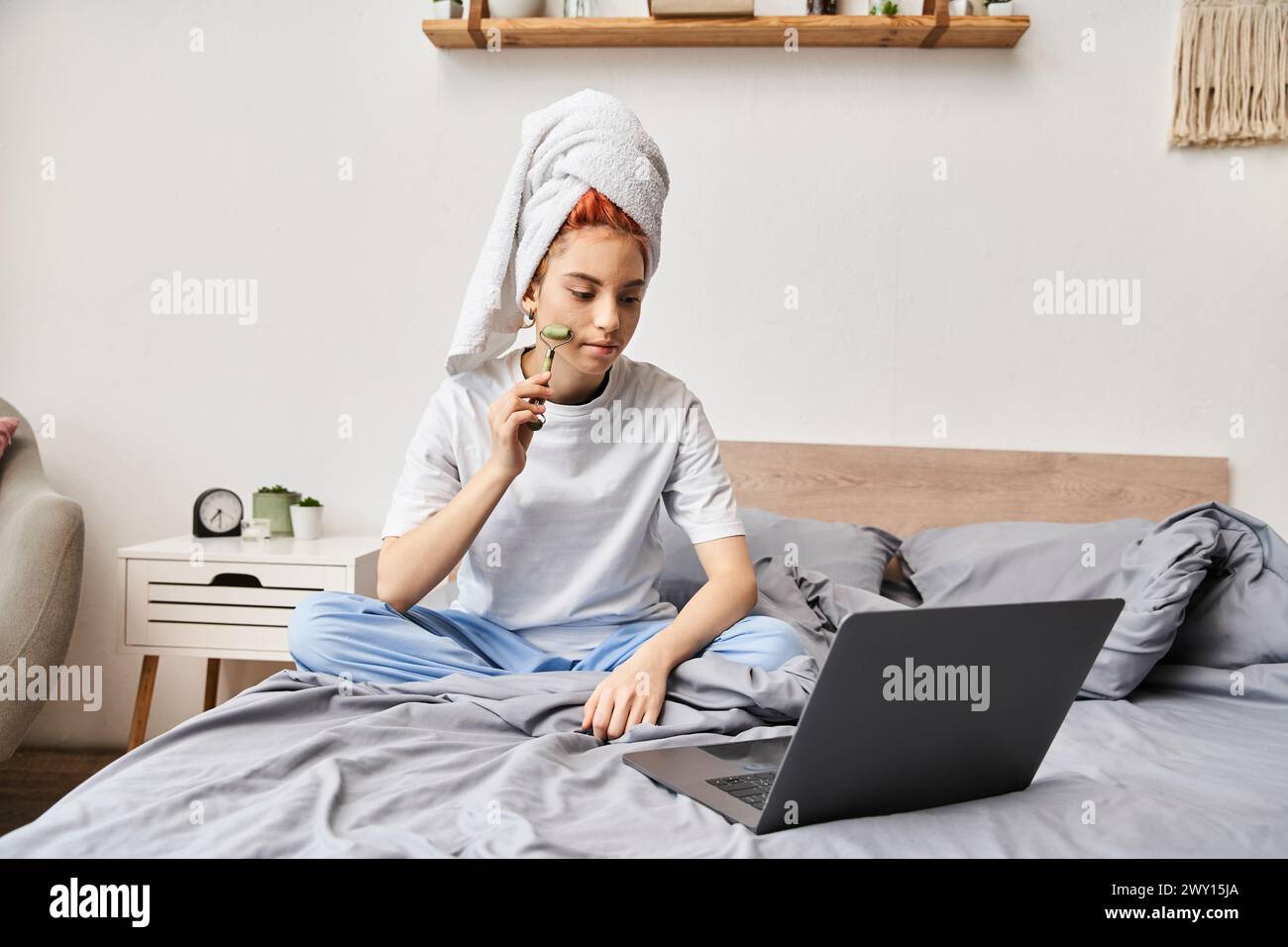 appealing red haired queer person in homewear using face roller while relaxing on bed with laptop Stock Photo
