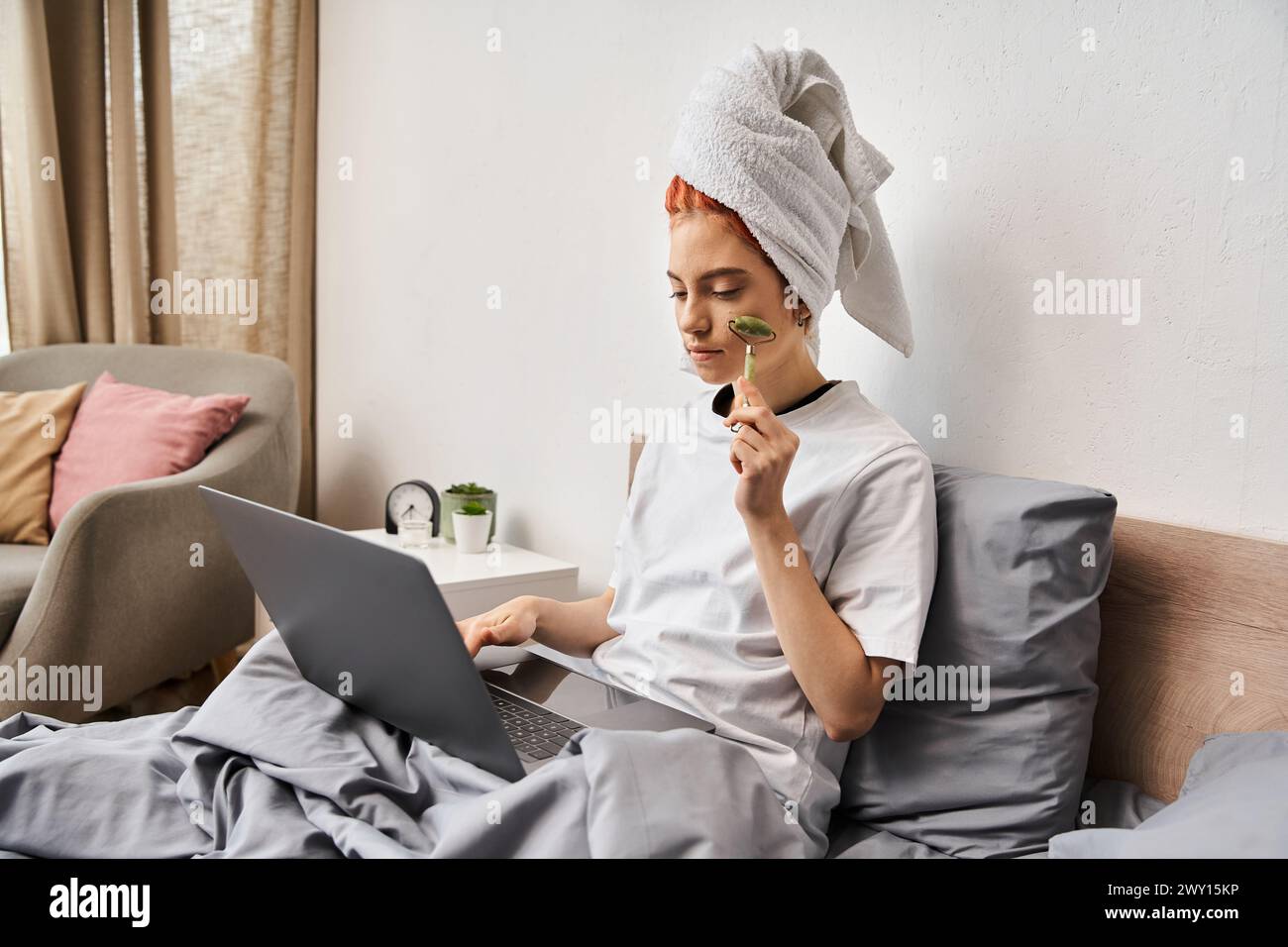 attractive red haired queer person in homewear using face roller while relaxing on bed with laptop Stock Photo